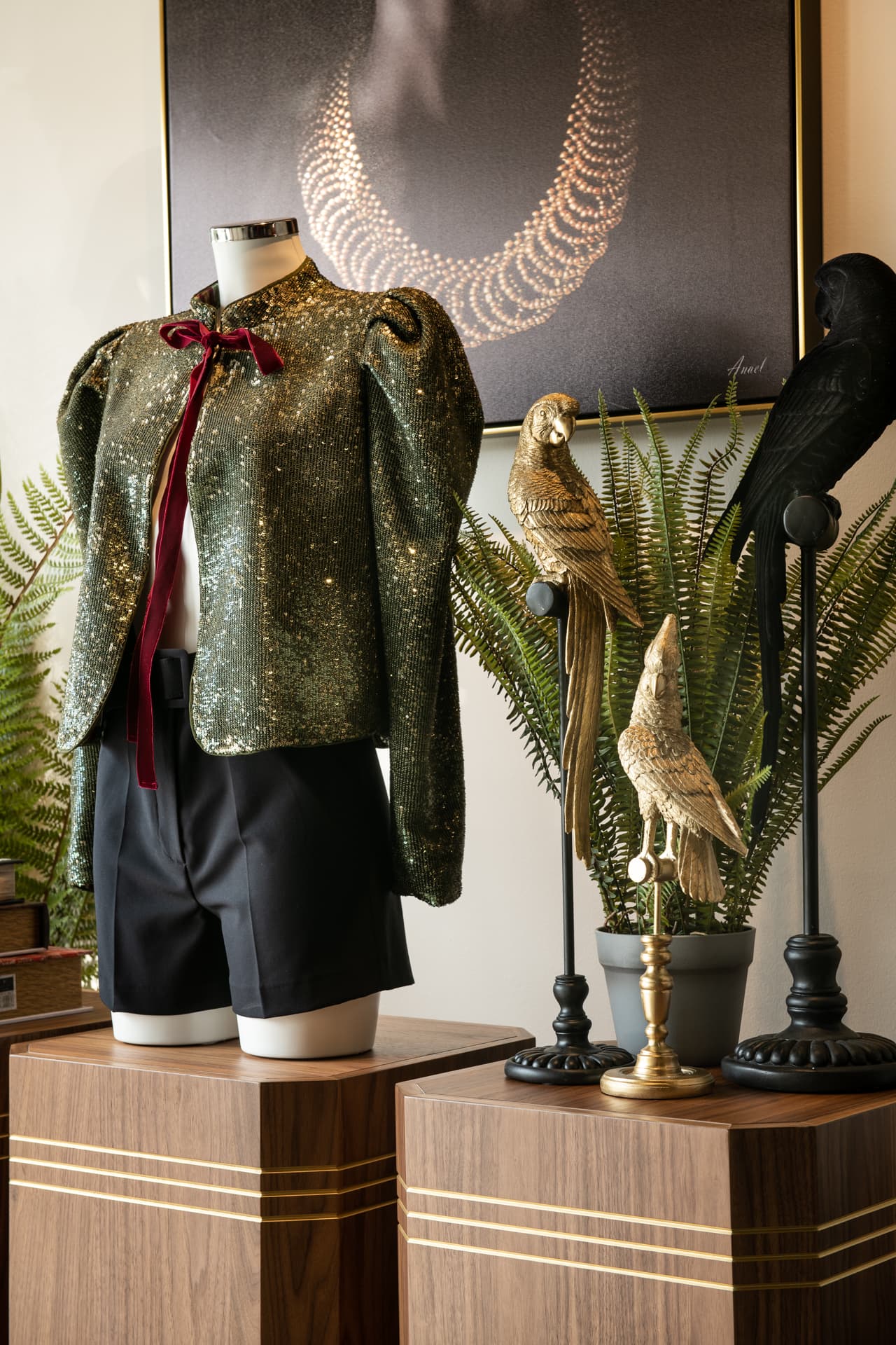 Sequinned jacket with a velvet bow on a bust form on a walnut plinth, beside gold and black parrot figurines, in the display area of DxVogue