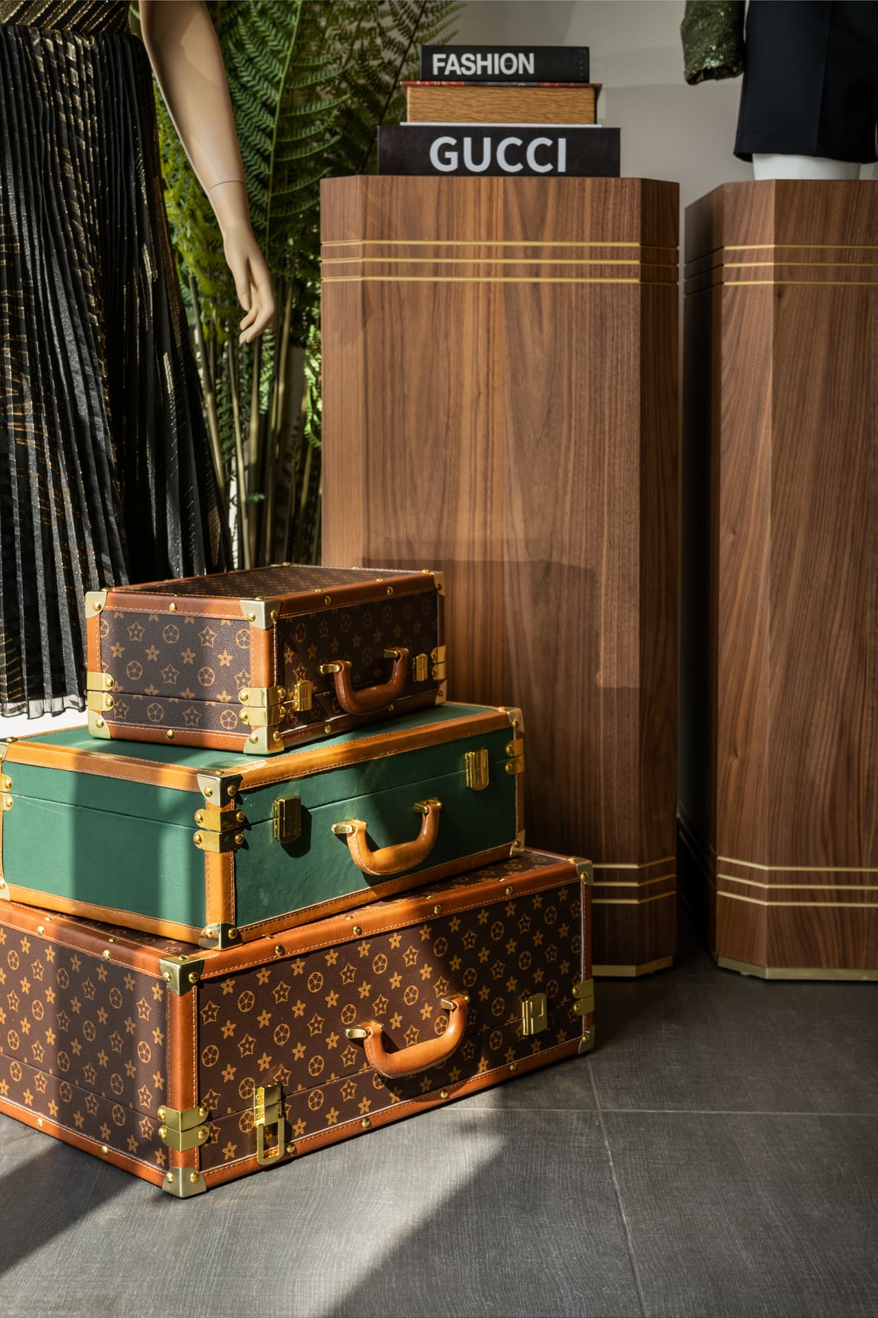 Three stacked vintage trunks with leather handles at the base of tall walnut plinths with brass bands, in the display area of DxVogue