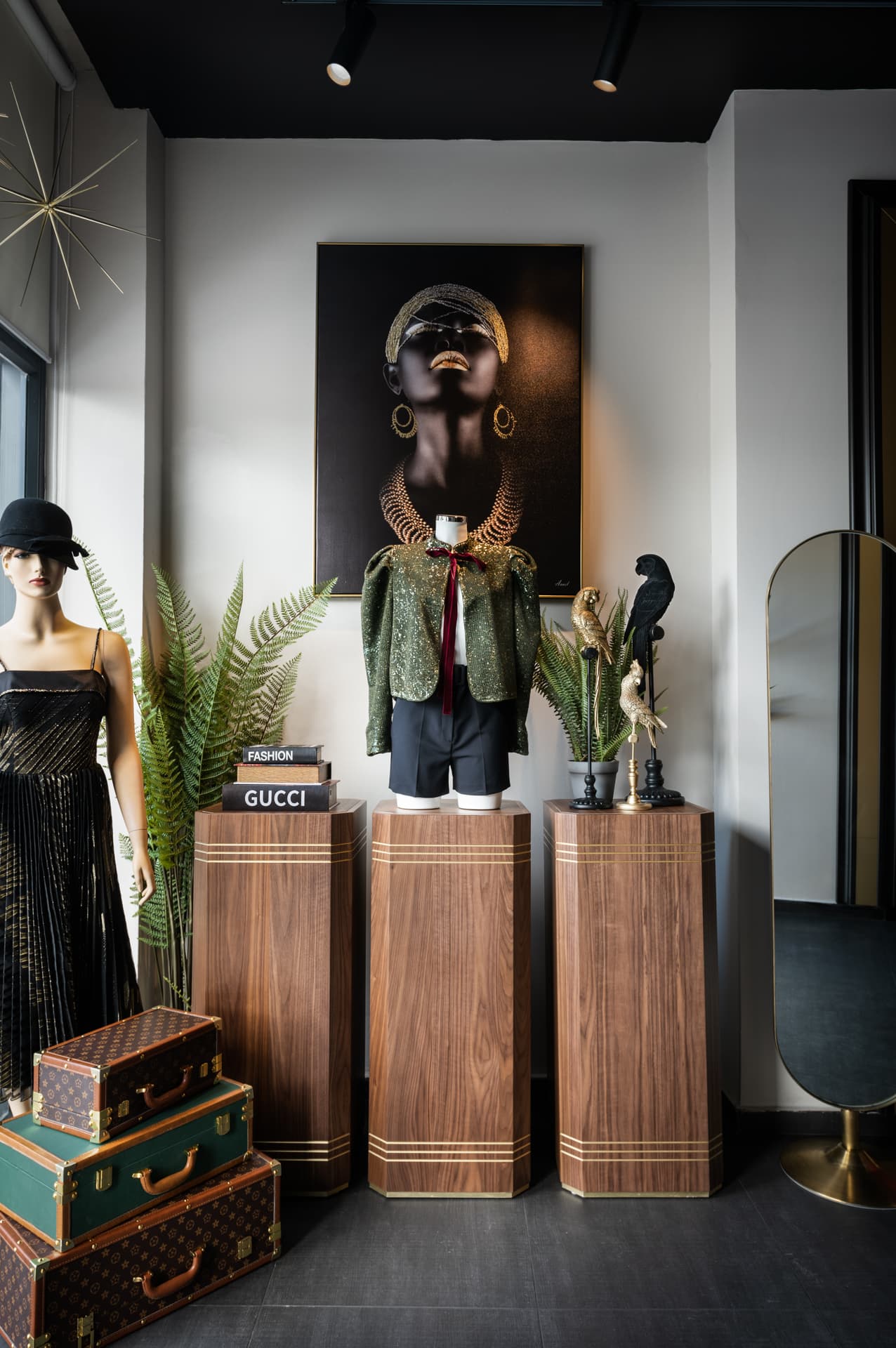 Three walnut plinths with brass inlay lines under a framed portrait, a mannequin and an oval floor mirror, in the display area of DxVogue