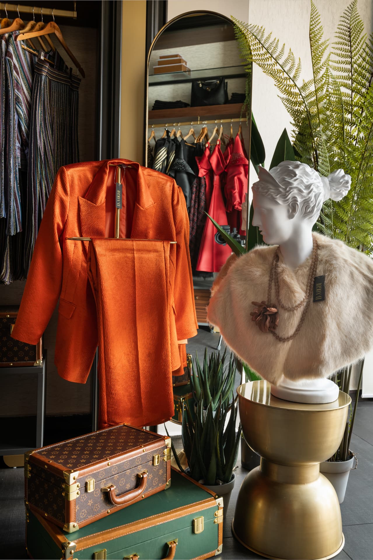 White classical bust in a fur stole on a brass side table beside an orange suit and stacked vintage trunks, in the display area of DxVogue