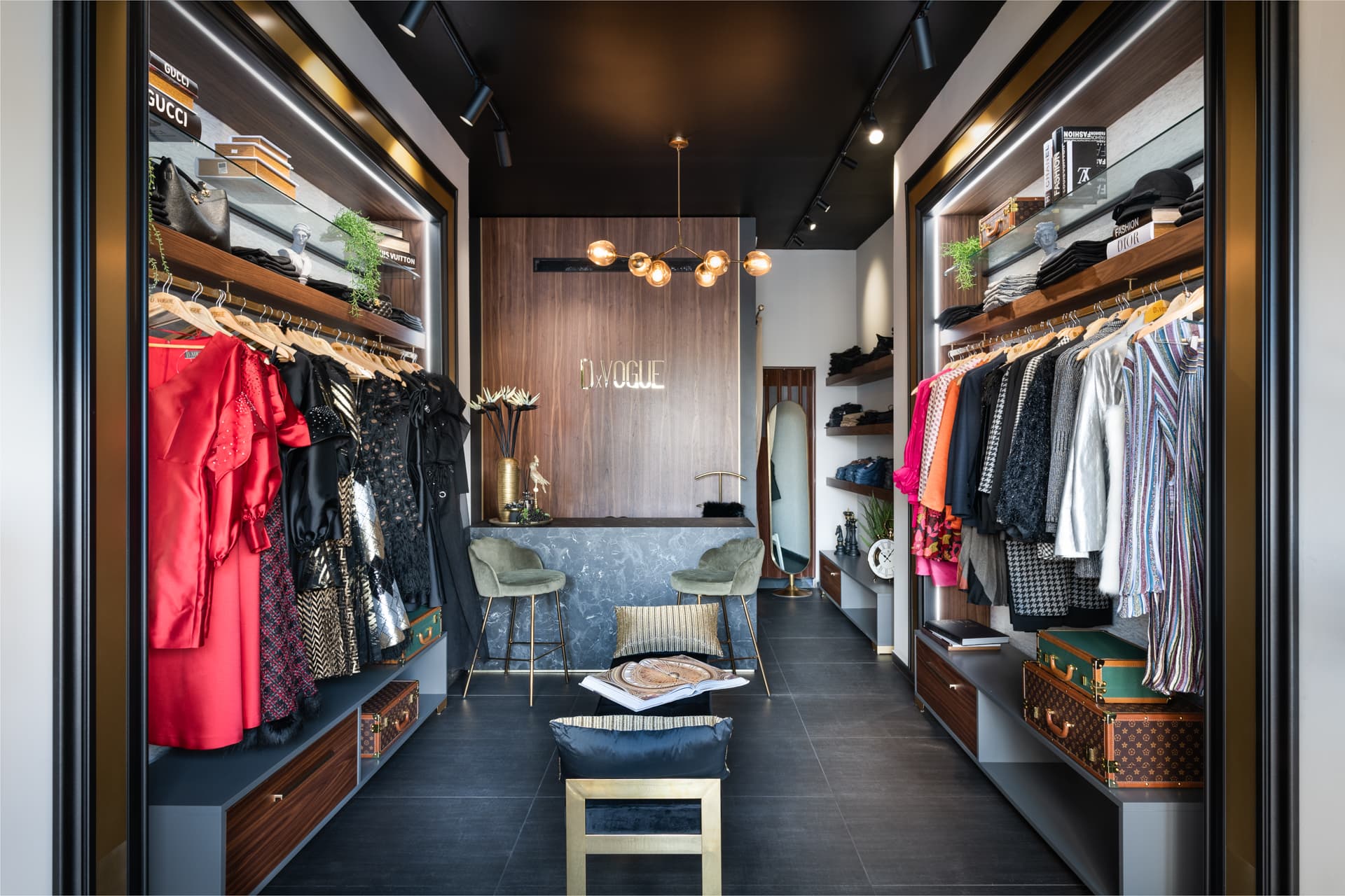 View down the boutique between lit walnut wardrobe bays toward a dark stone counter and globe chandelier, in the showroom of DxVogue