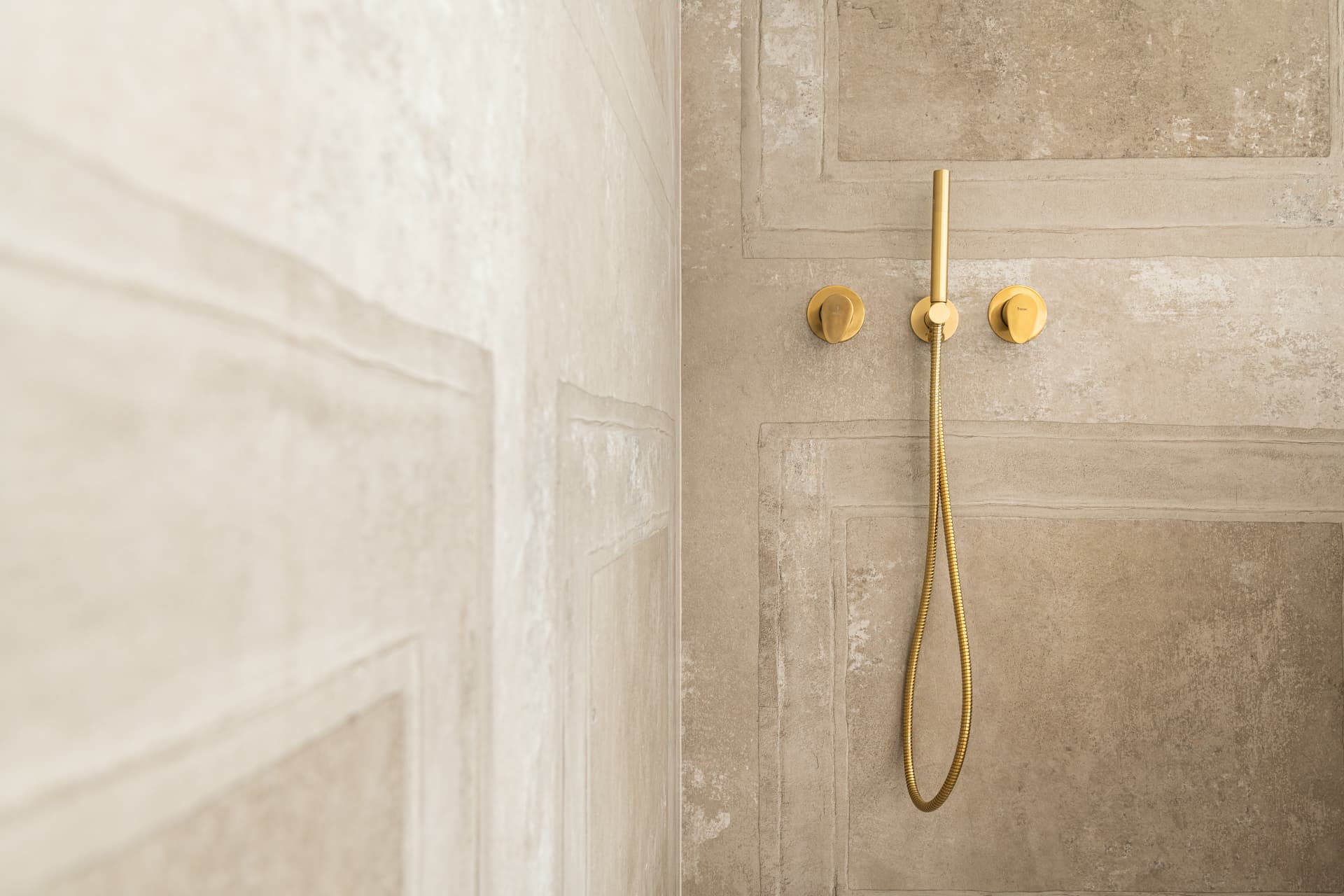 Brass hand shower on a hose between two round brass valves on a panel-molded beige shower wall in the bathroom of Emperador Dark