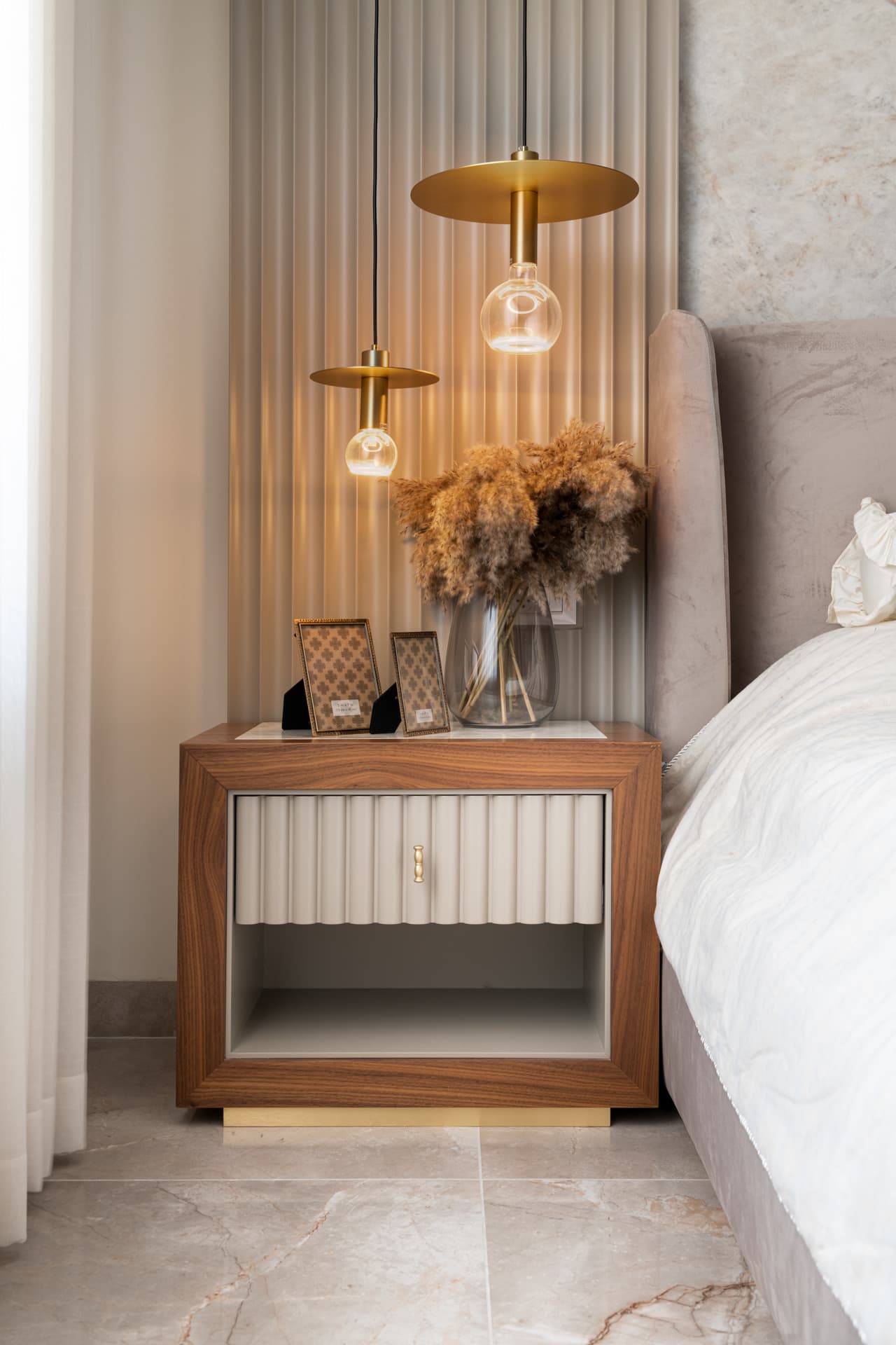 Walnut nightstand with a fluted drawer beneath brass disc pendant lights beside the bed in the master bedroom of Emperador Dark