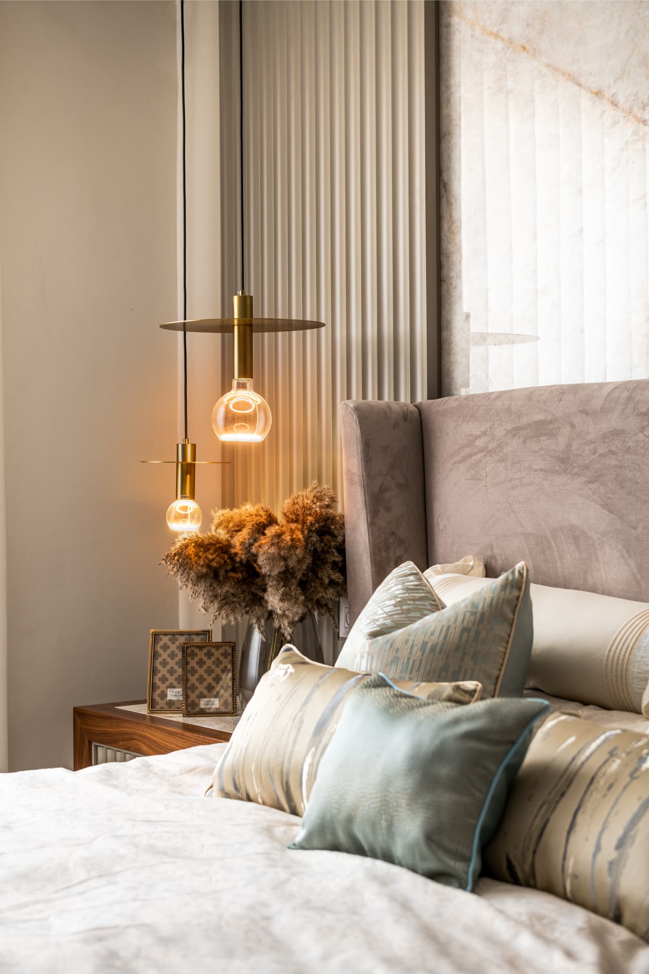 Glowing brass disc pendants beside a grey velvet wingback headboard and patterned cushions in the master bedroom of Emperador Dark