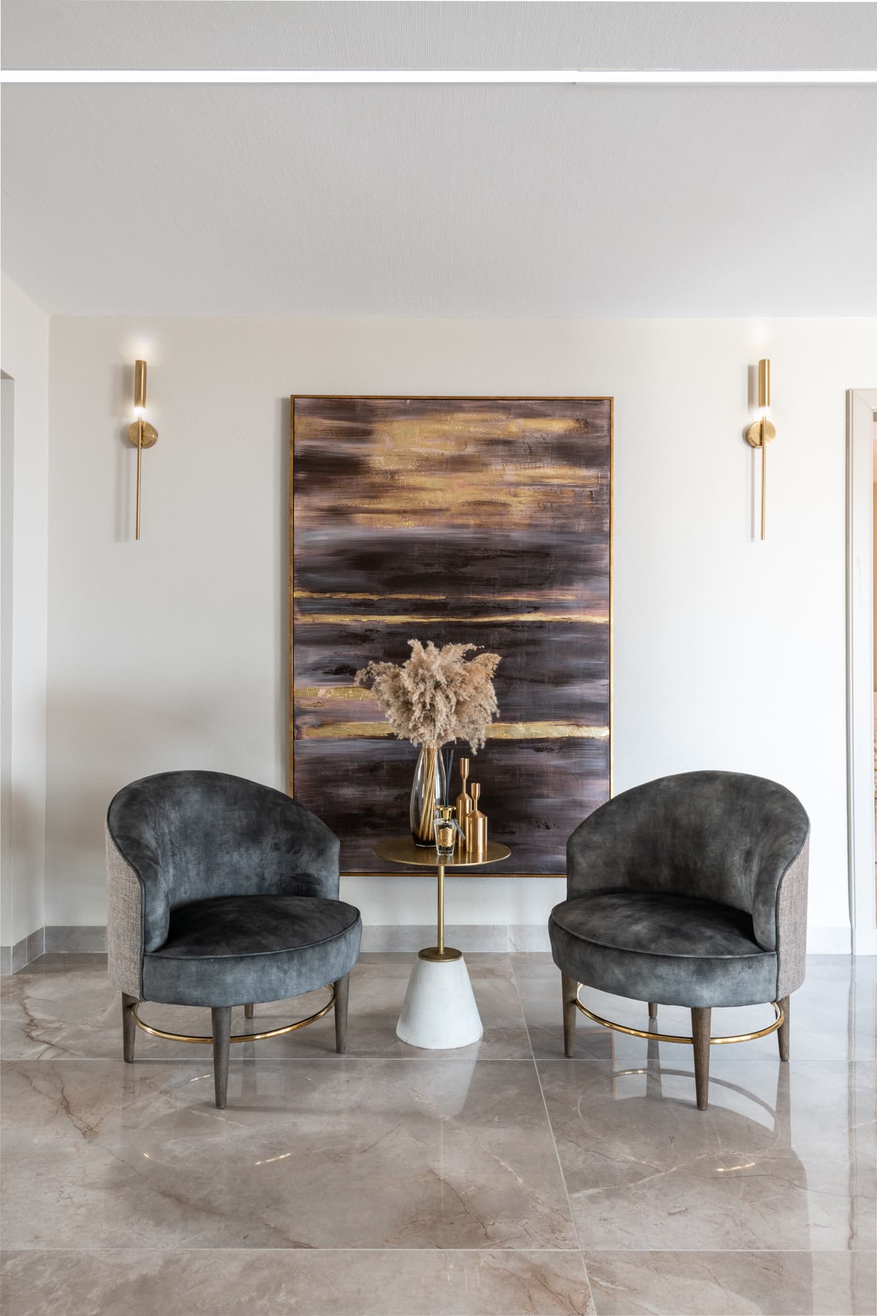Two grey velvet tub chairs flank a brass-topped side table beneath a gold-streaked abstract painting in the entrance of Emperador Dark