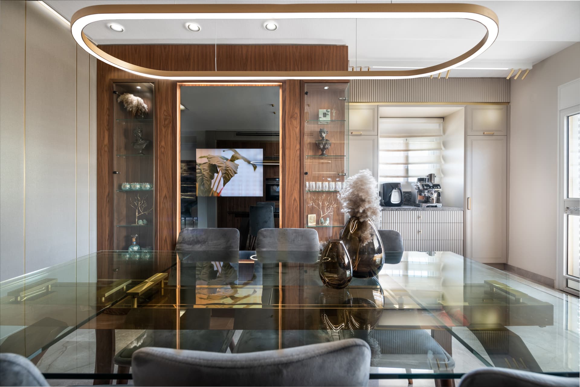 Glass dining table under an oval brass ring pendant, facing lit walnut display cabinets in the kitchen of Emperador Dark