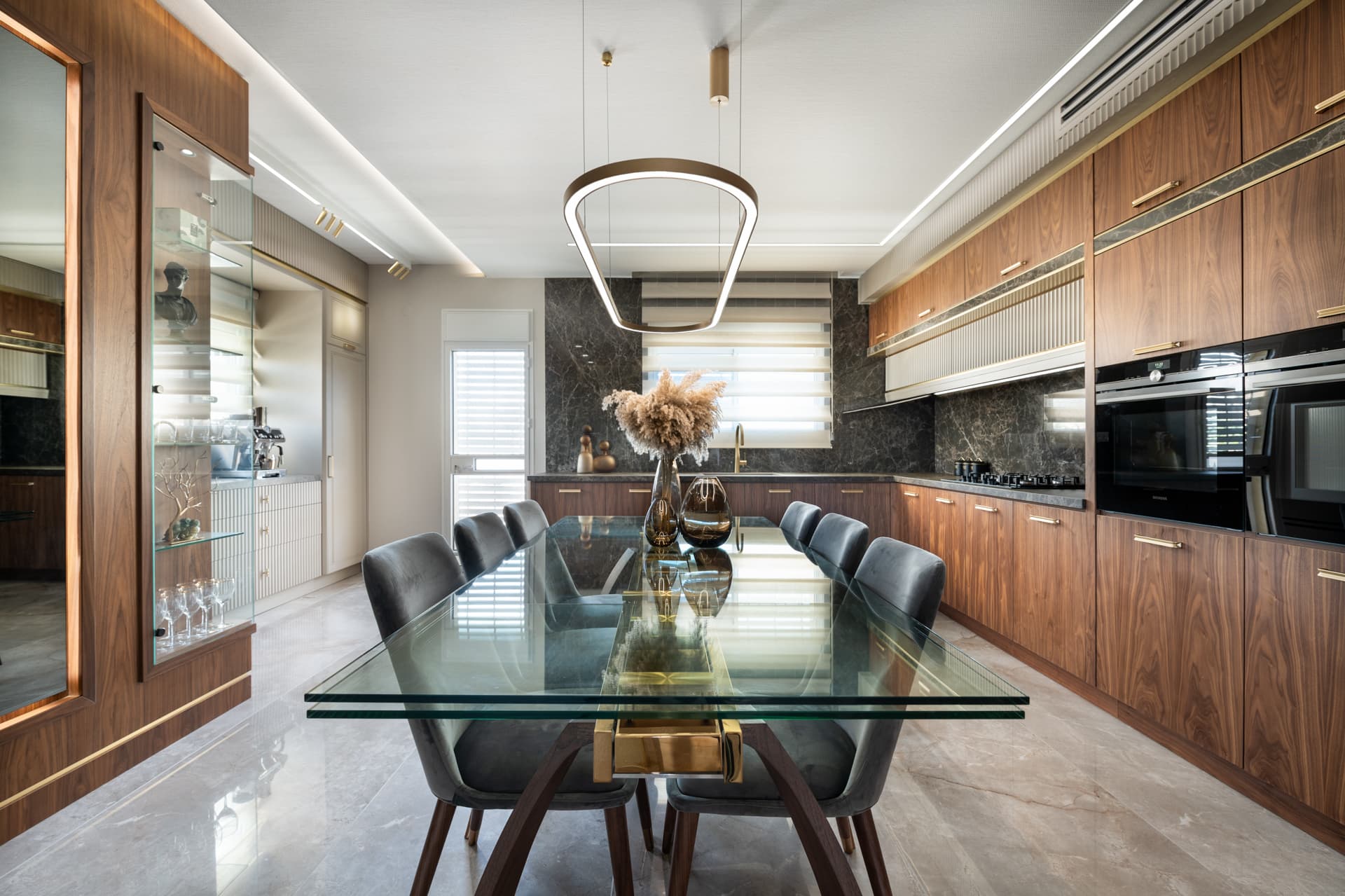 Long glass dining table with grey velvet chairs beside walnut cabinetry and a dark veined stone backsplash in the kitchen of Emperador Dark