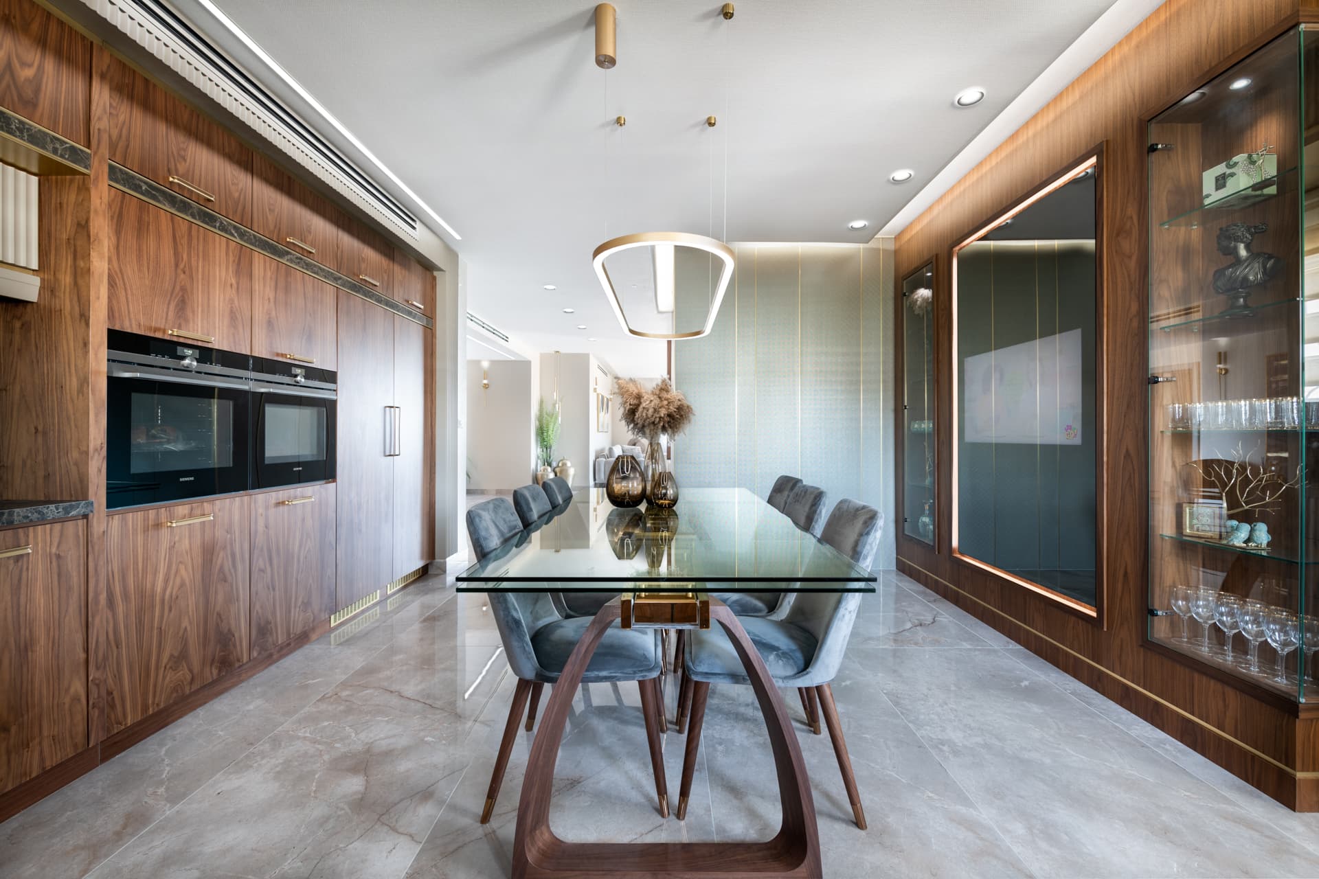 View along the glass dining table between built-in ovens and illuminated walnut glass cabinets in the kitchen of Emperador Dark