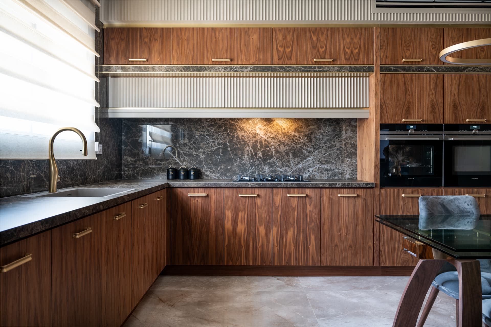 Corner of walnut base cabinets with a brass faucet and a lit dark veined stone backsplash in the kitchen of Emperador Dark