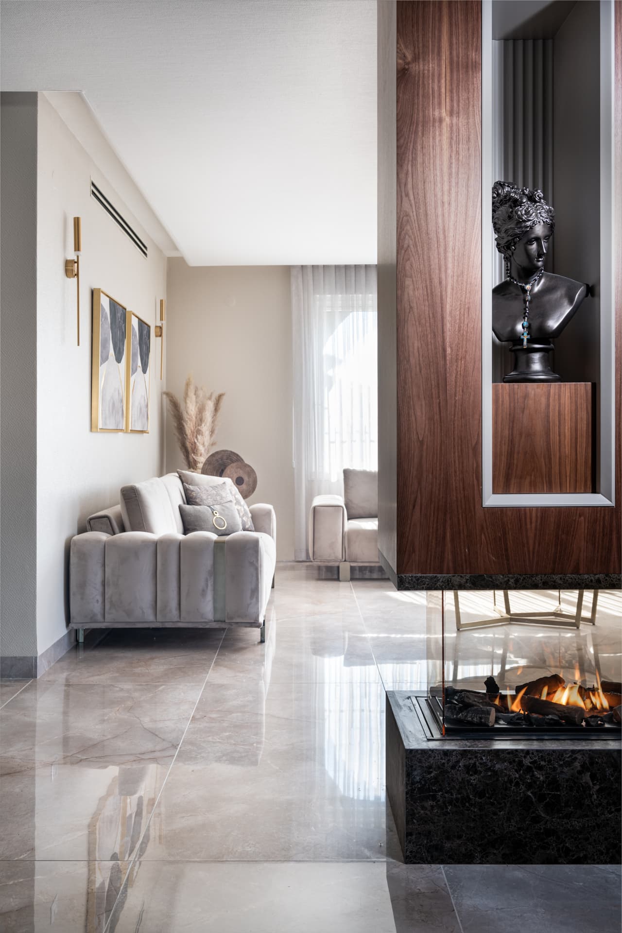 Black sculpted bust in a walnut niche above a glass-sided fireplace, with a tufted grey sofa beyond, in the living room of Emperador Dark