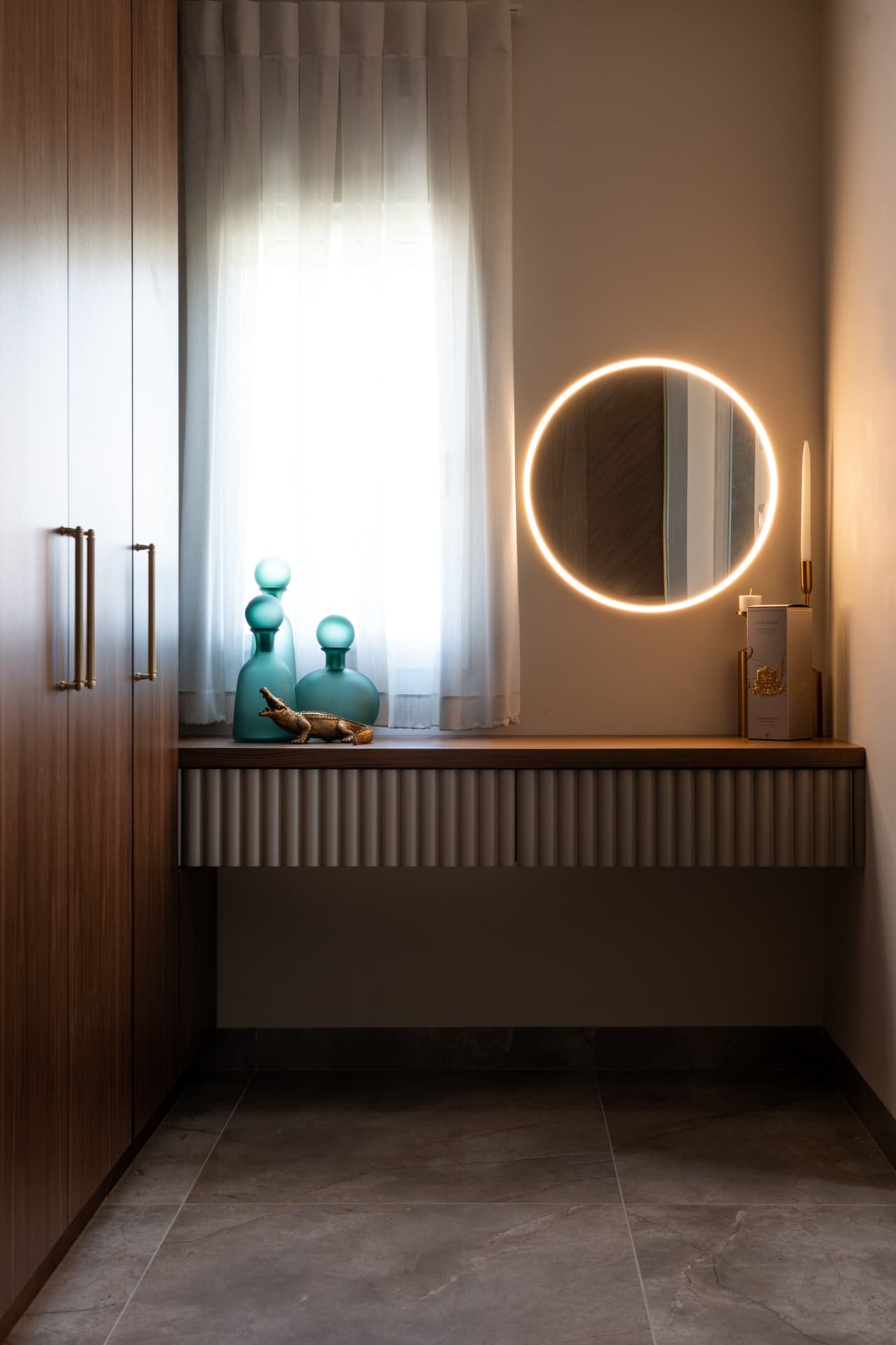 Floating fluted vanity with a backlit round mirror and green glass bottles below a window in the master bedroom of Emperador Dark