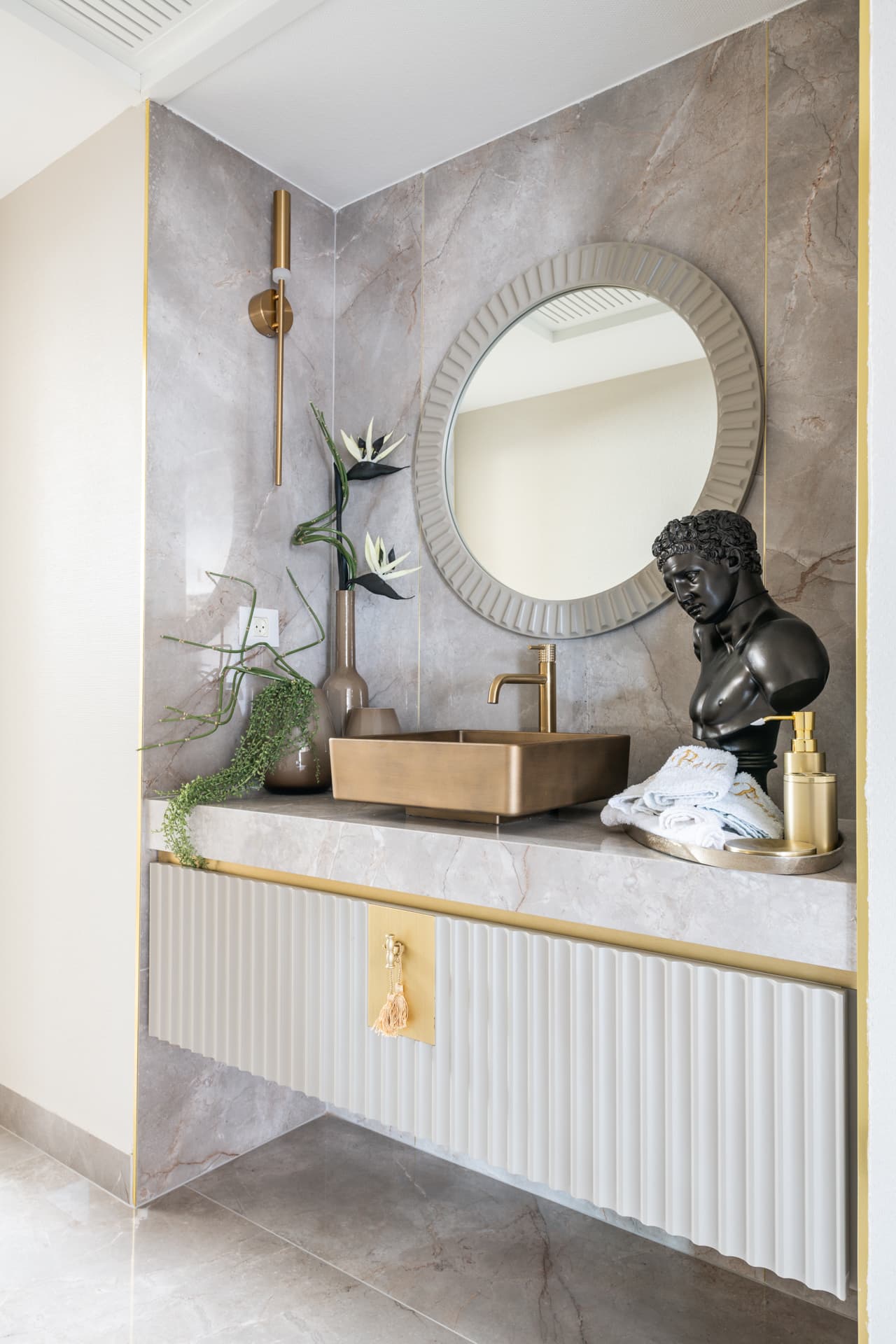 Brass vessel sink on a stone counter over a fluted white vanity with a black bust and ribbed mirror in the powder room of Emperador Dark