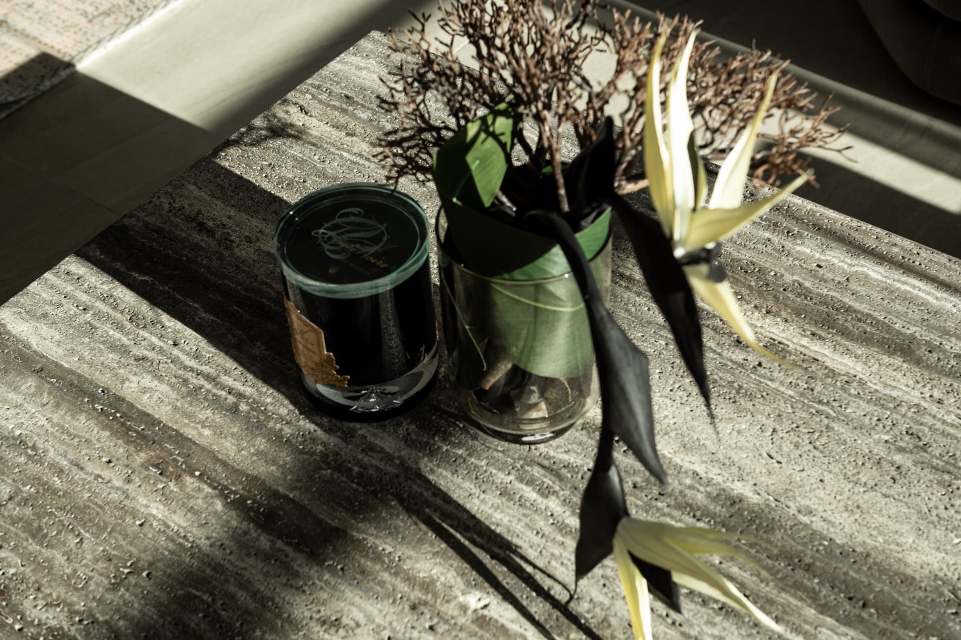 Glass vase with dried branches and a leaf beside a black candle jar on a veined stone tabletop in raking sunlight in Khayyal