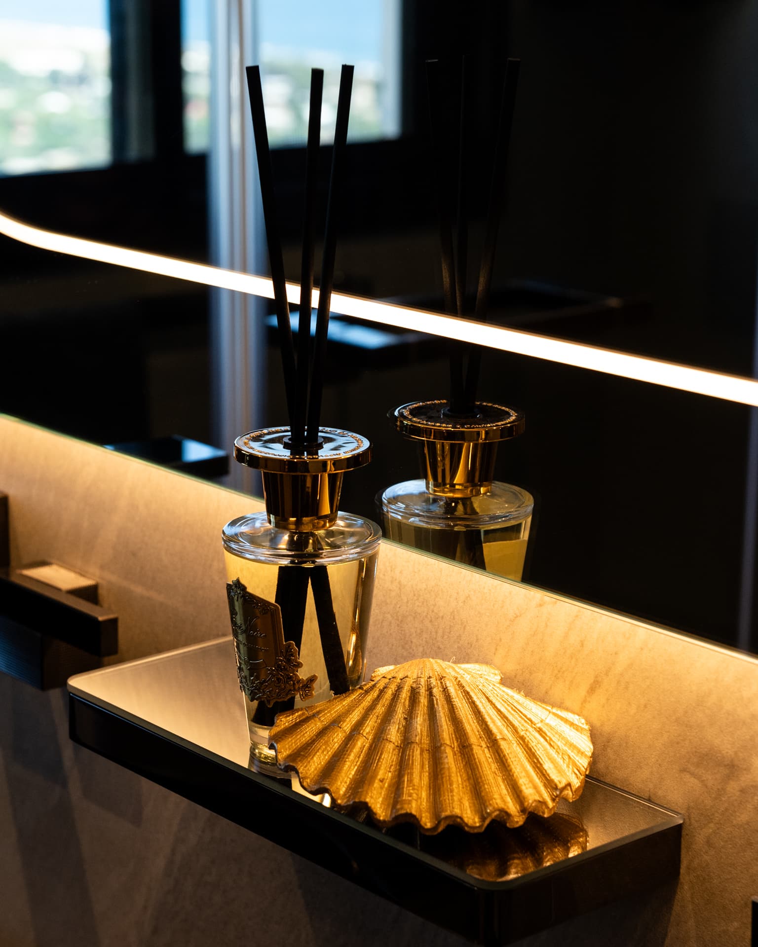 Gold scallop shell and reed diffusers on a black glass shelf beneath a backlit mirror in Khayyal