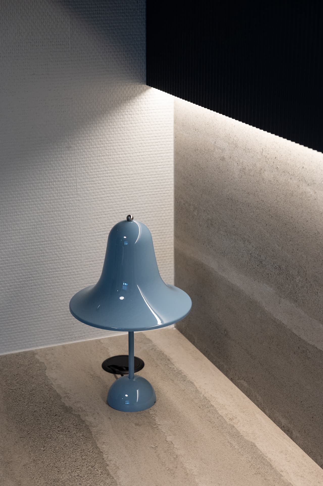 Blue bell-shaped table lamp on a beige stone desk under a lit shelf with a textured wall in a kids room of Khayyal