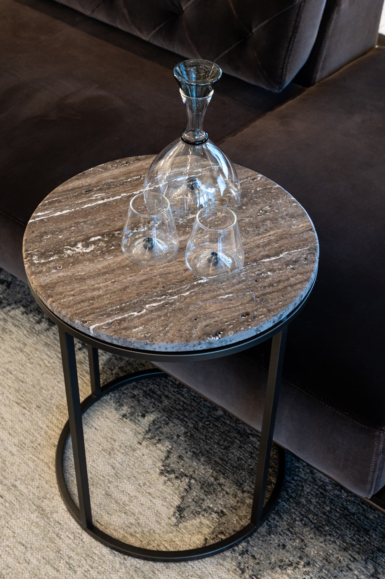 Round veined stone side table on a black metal frame with a glass carafe set beside the tufted sofa in the salon of Khayyal