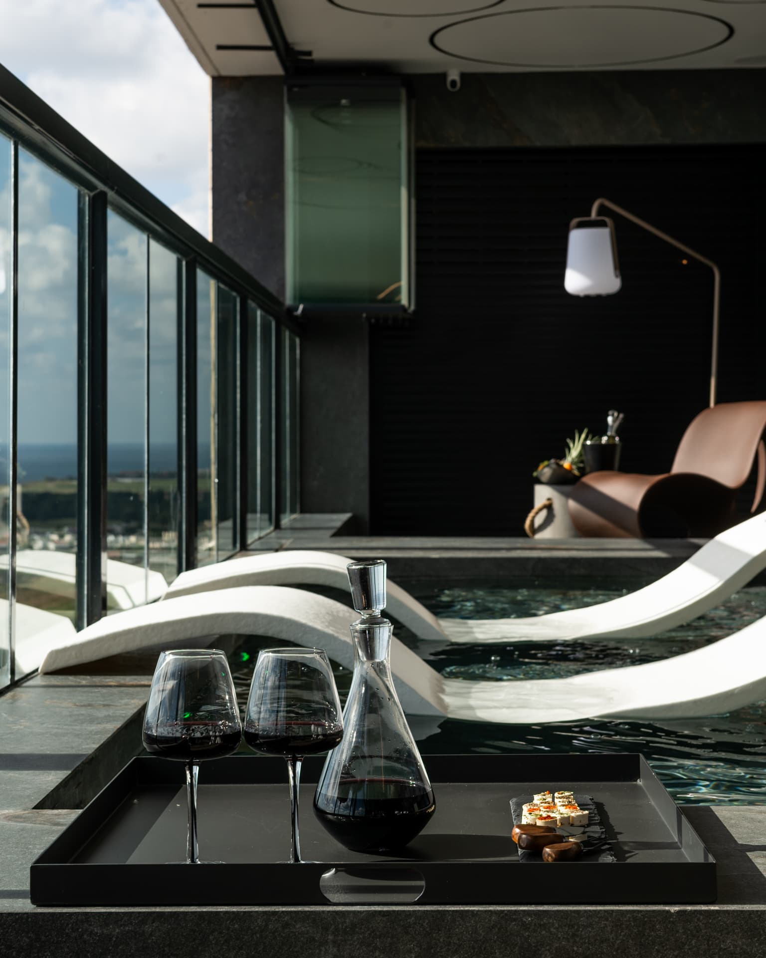 Black tray with red wine glasses, a decanter and small bites on the pool edge beside white loungers in Khayyal