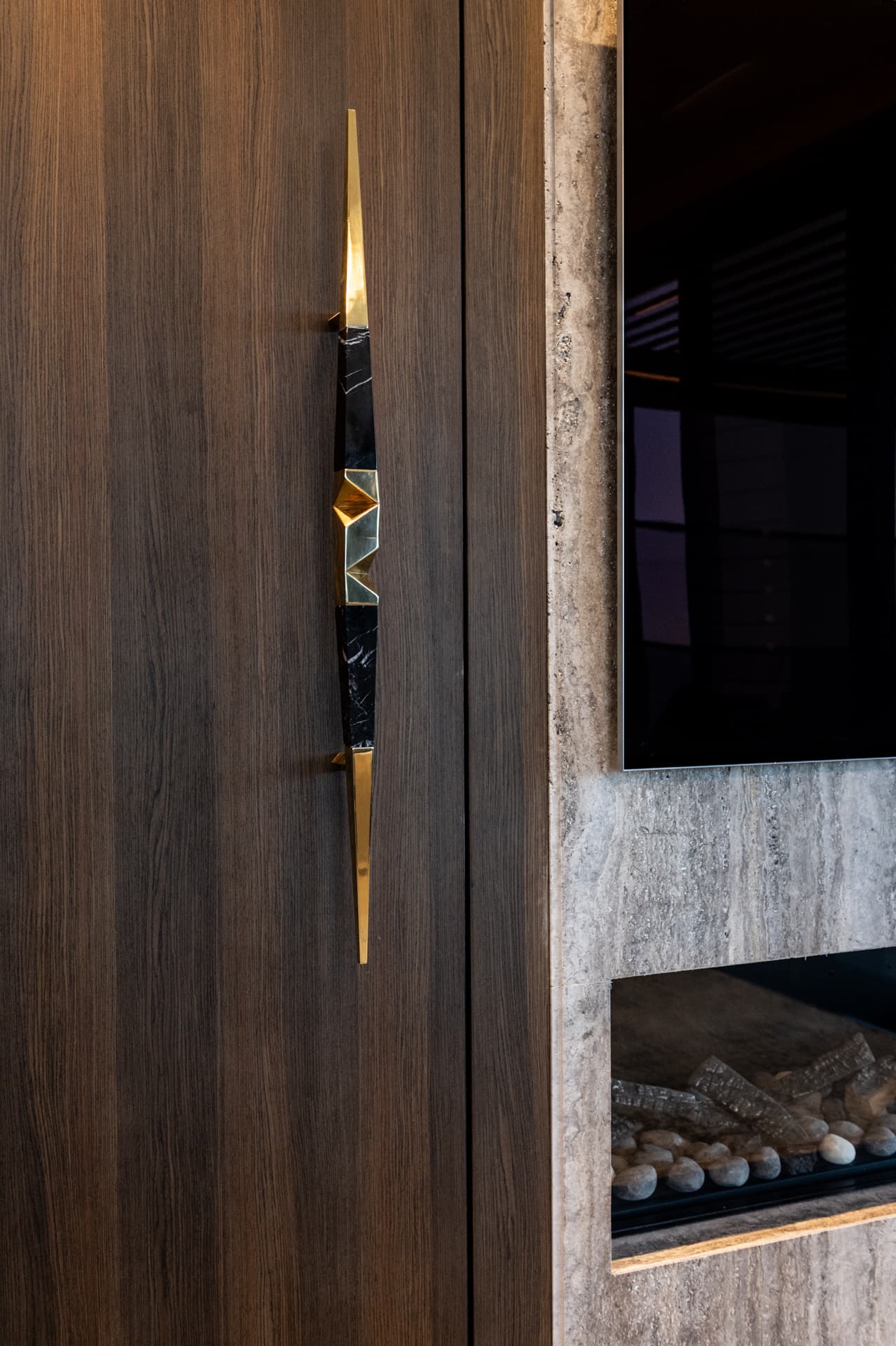 Long brass and black stone door pull on a walnut door beside the stone fireplace wall in the living room of Khayyal