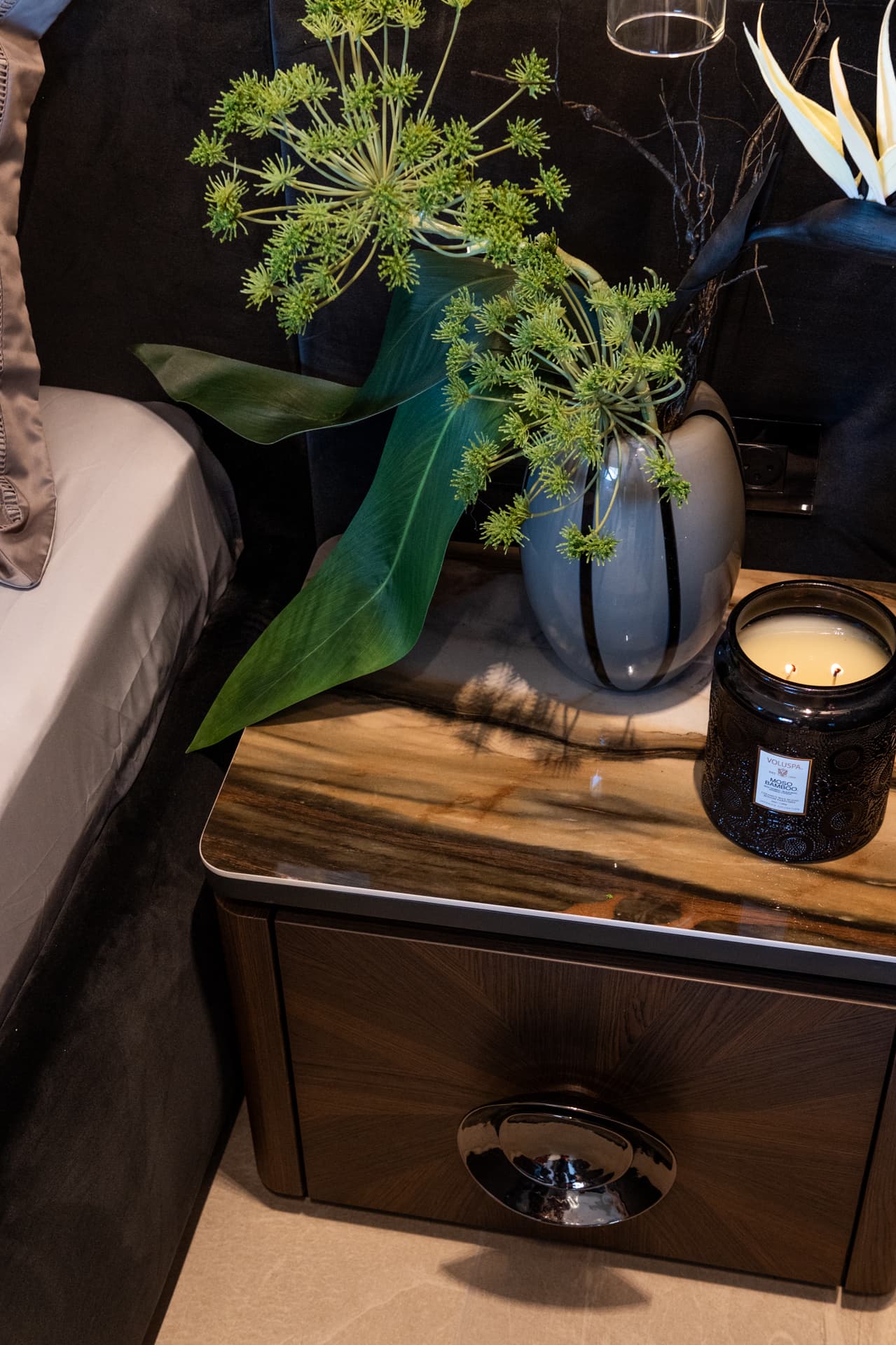 Sunburst-veneer nightstand with a glossy dark round pull, polished top, ceramic vase and candle jar in the master bedroom of Khayyal