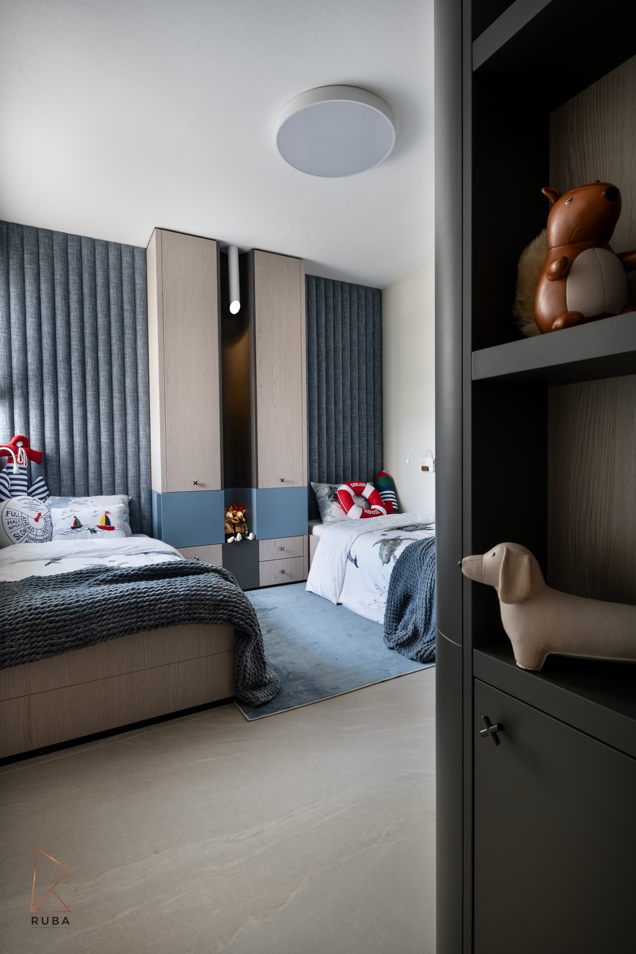 Twin-bed kids room with a blue channel-tufted wall seen past a dark display shelf of toy animals in Khayyal