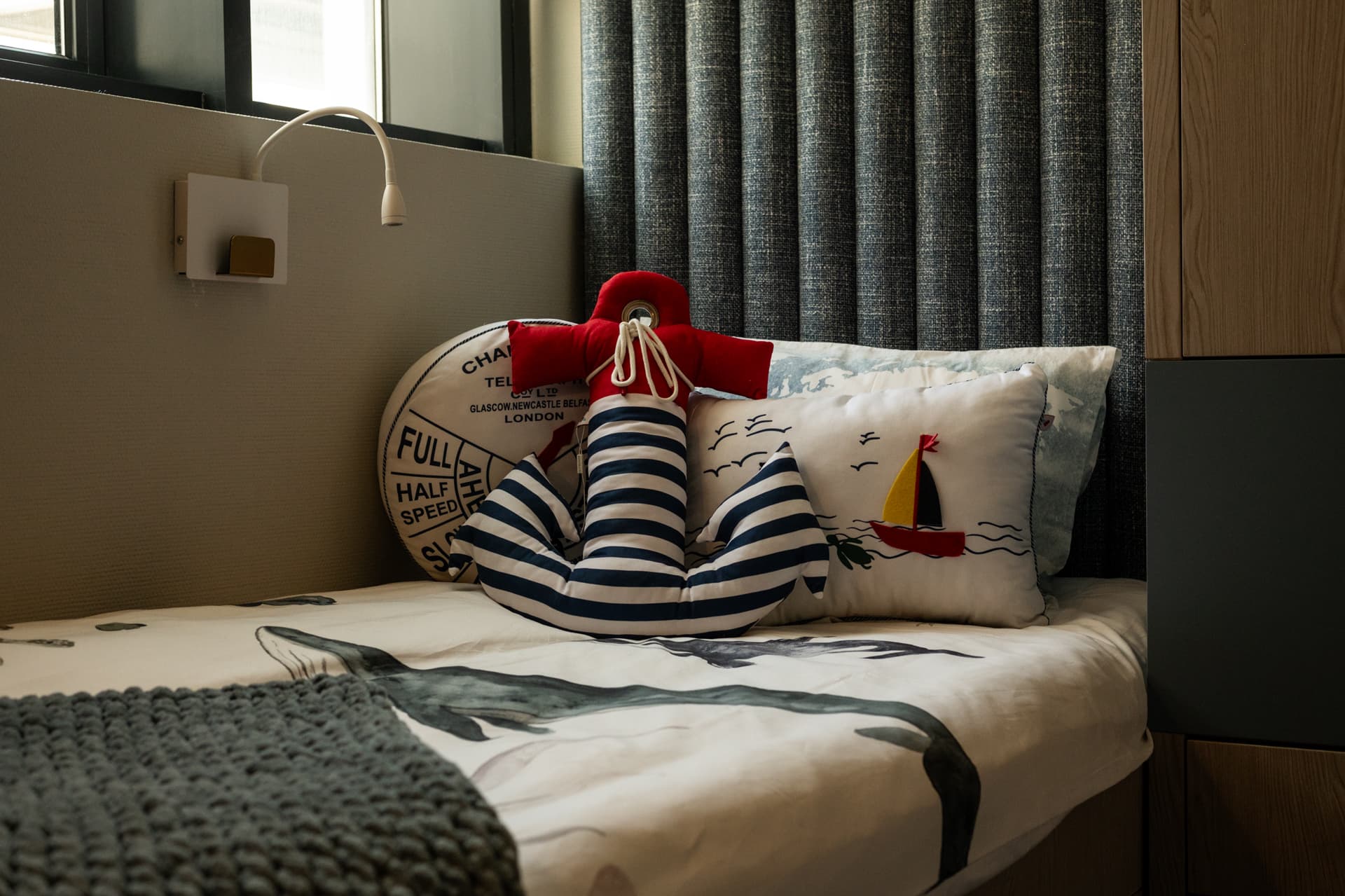 Anchor-shaped cushion and sailboat pillow on a bed with a wall reading lamp and channel-upholstered wall in a kids room of Khayyal