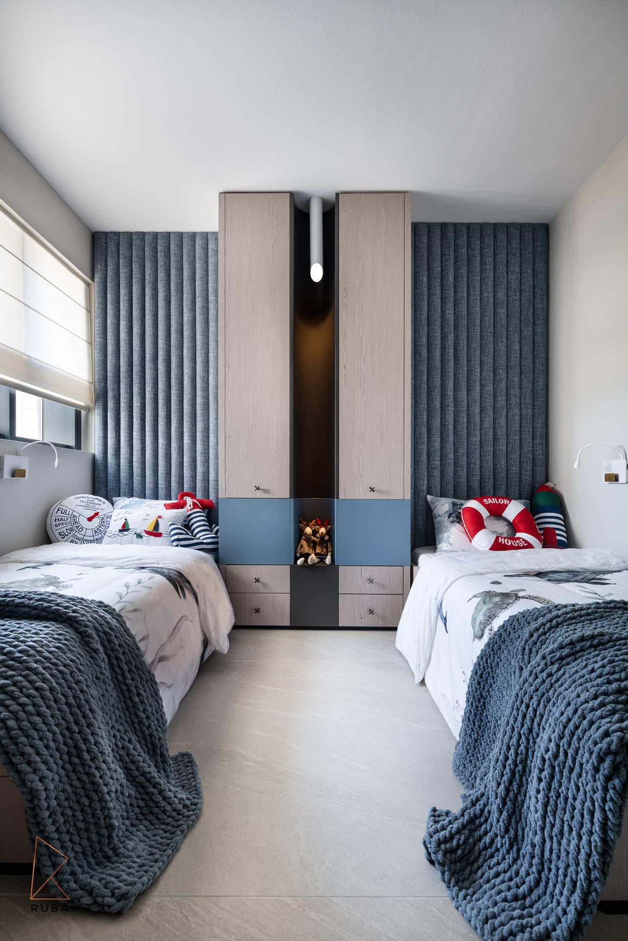 Twin beds flanking a central wood cabinet on a blue channel-tufted wall with knitted throws in a kids room of Khayyal
