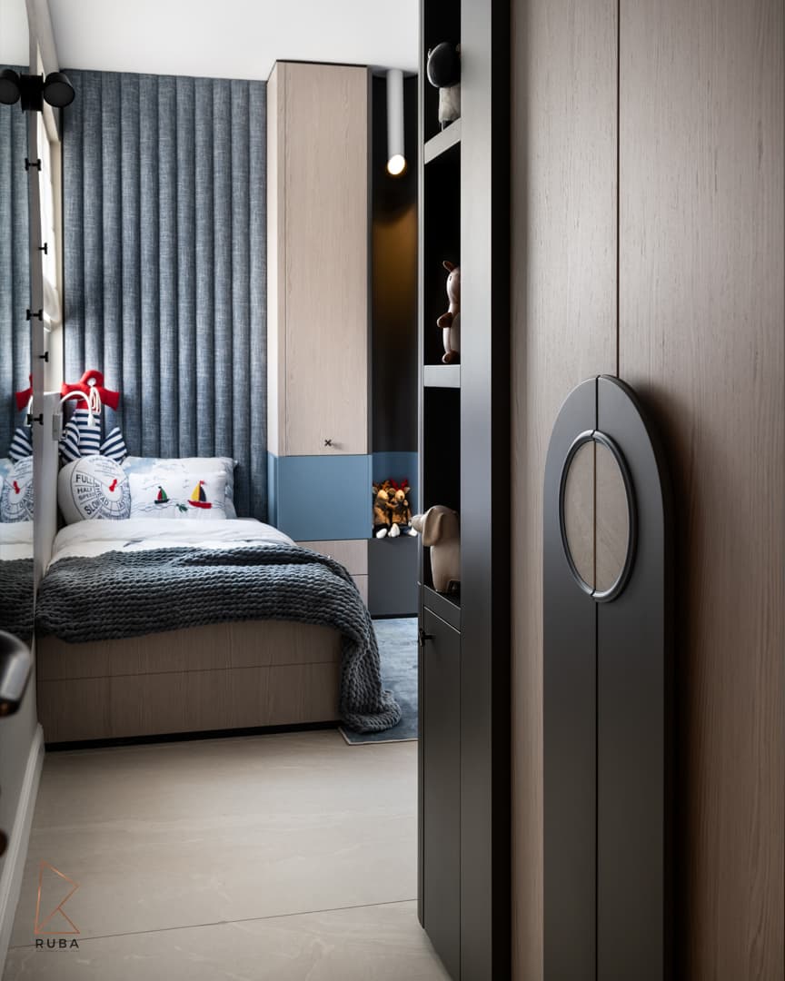 Pale wood wardrobe door with a dark arched panel and round pull framing a view of a bed and blue tufted wall in a kids room of Khayyal