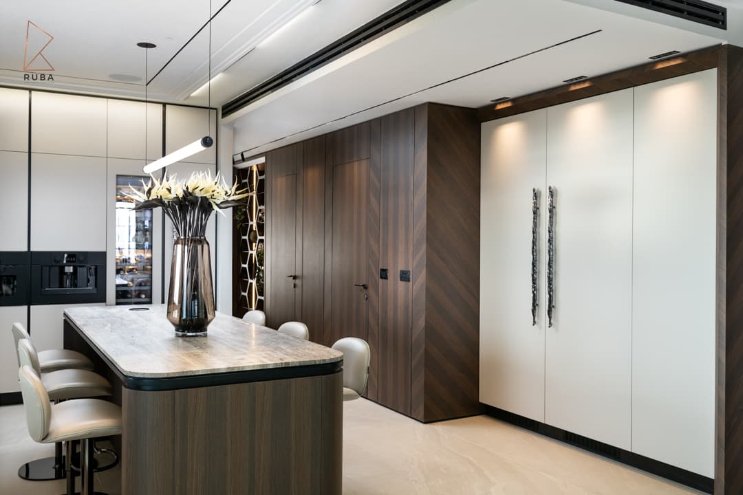 Kitchen island with a veined stone top and bar stools between walnut chevron doors and tall white cabinets in Khayyal