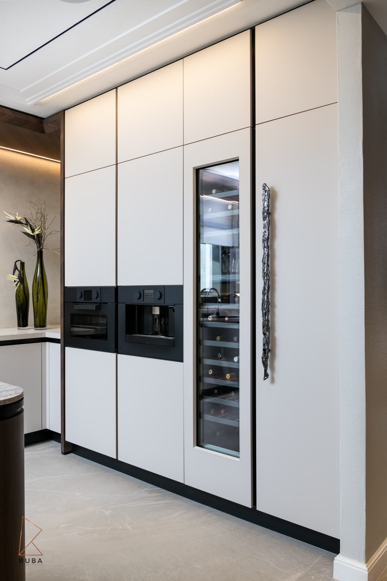 Tall white cabinetry with built-in ovens, a glass-front wine fridge and a sculpted long pull in the kitchen of Khayyal