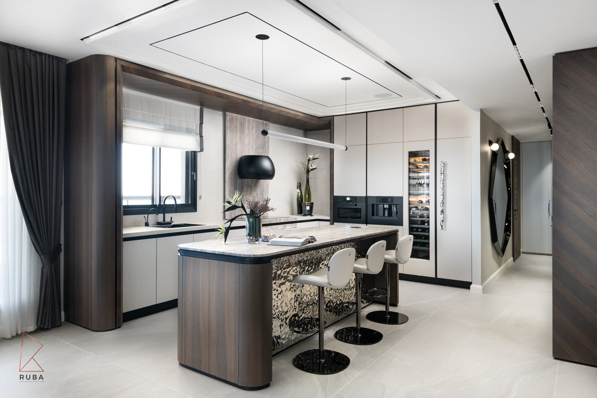Open kitchen with a rounded walnut island, white bar stools, black dome hood and a tall wine fridge leading to a hallway in Khayyal