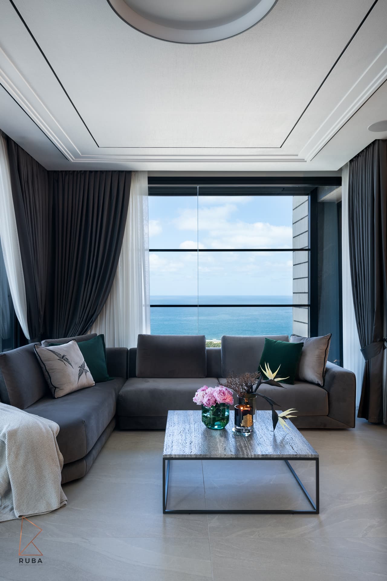 Grey sectional sofa facing a floor-to-ceiling sea-view window with dark drapery and a stone coffee table in the living room of Khayyal
