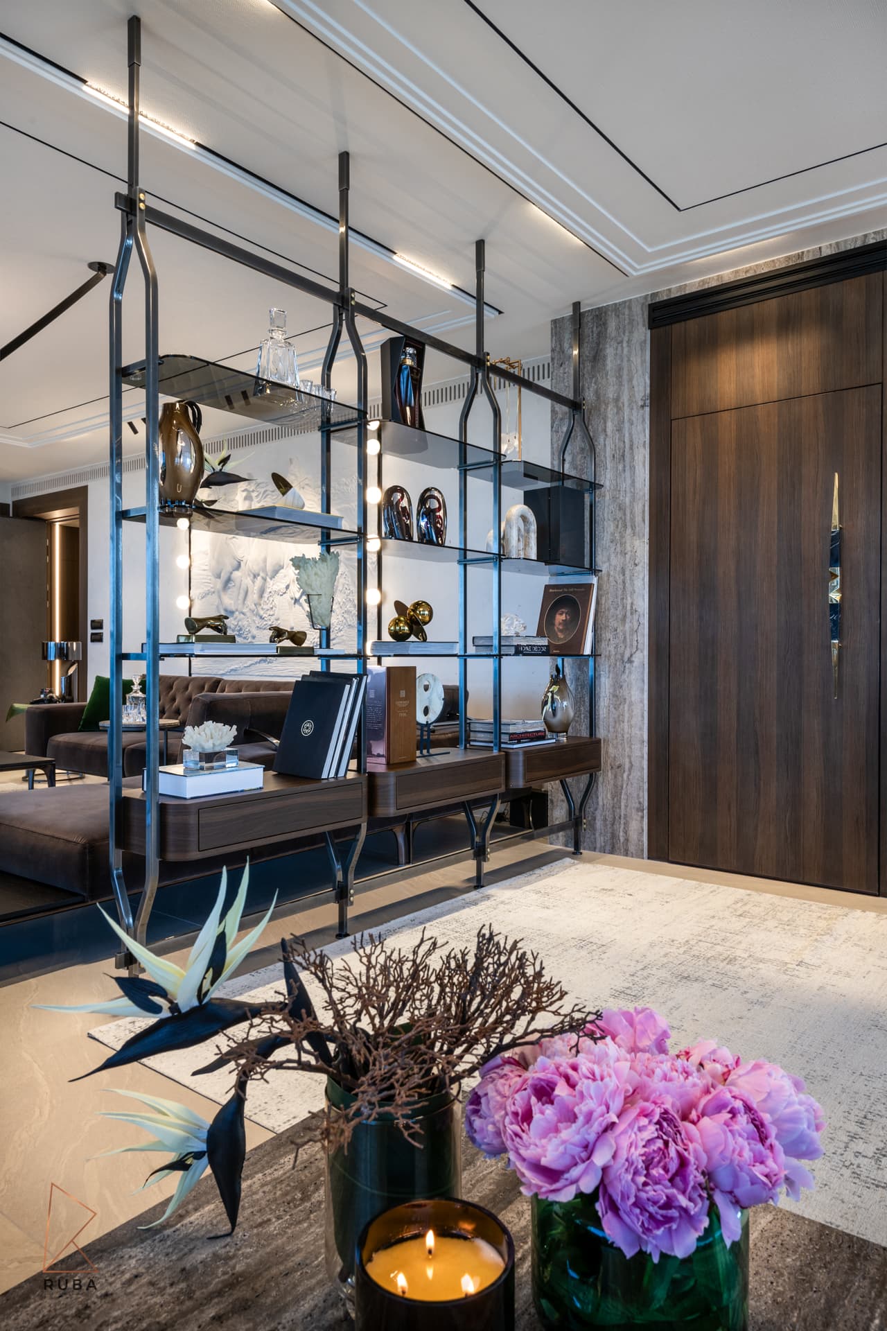 Close angle on the suspended glass shelving with its shelf lights on, peonies in front and a sofa beyond in the living room of Khayyal