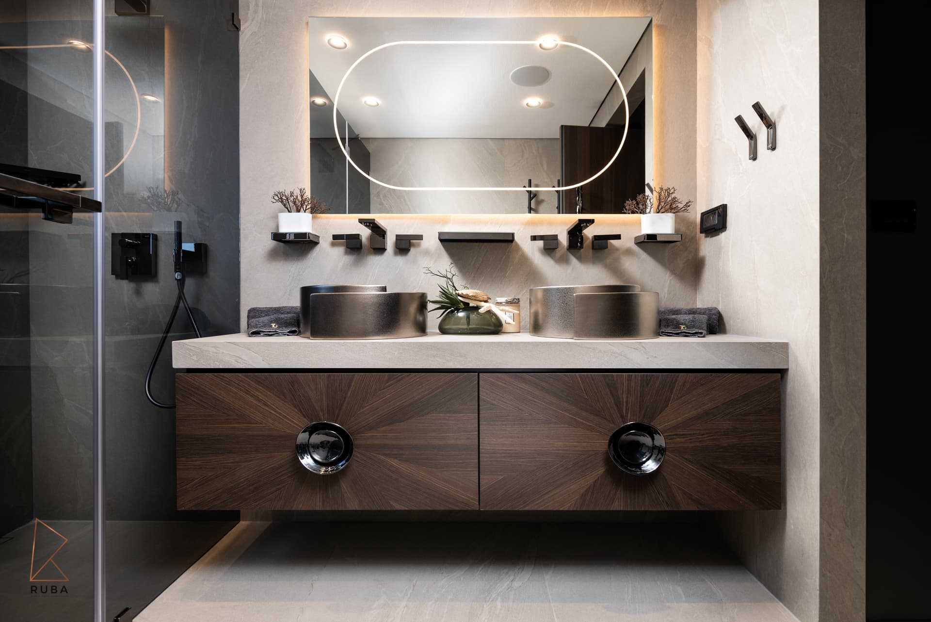 Double vanity in sunburst walnut veneer with round metallic basins under a mirror with an oval light ring in the master bathroom of Khayyal