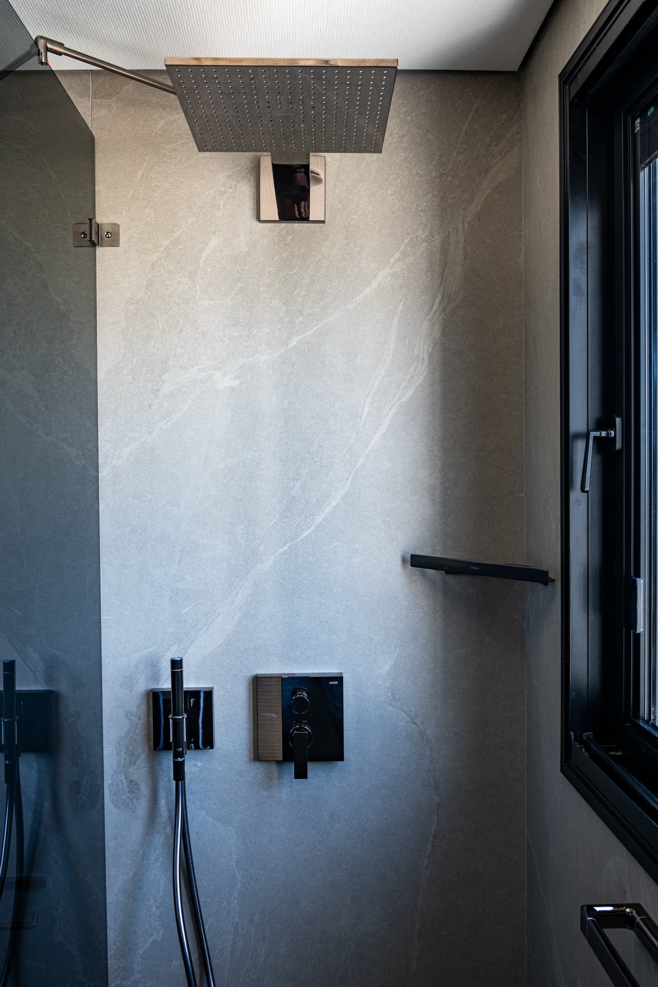 Walk-in shower with a square rain head, black mixer and stone-look walls beside a tall window in the master bathroom of Khayyal