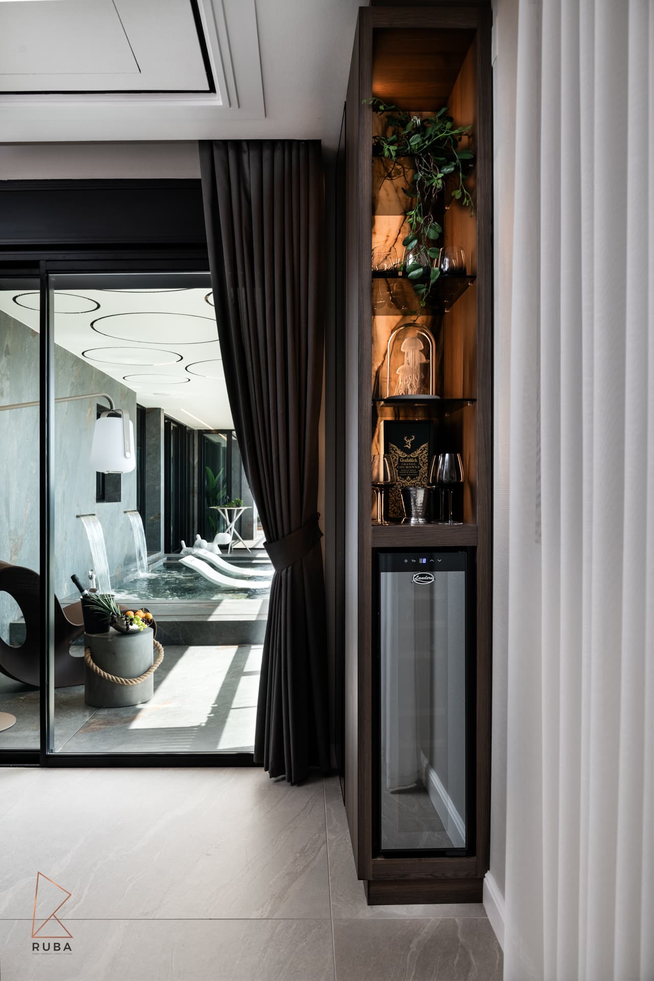 Backlit walnut display niche with a built-in wine fridge beside curtains opening onto the pool terrace from the master bedroom of Khayyal