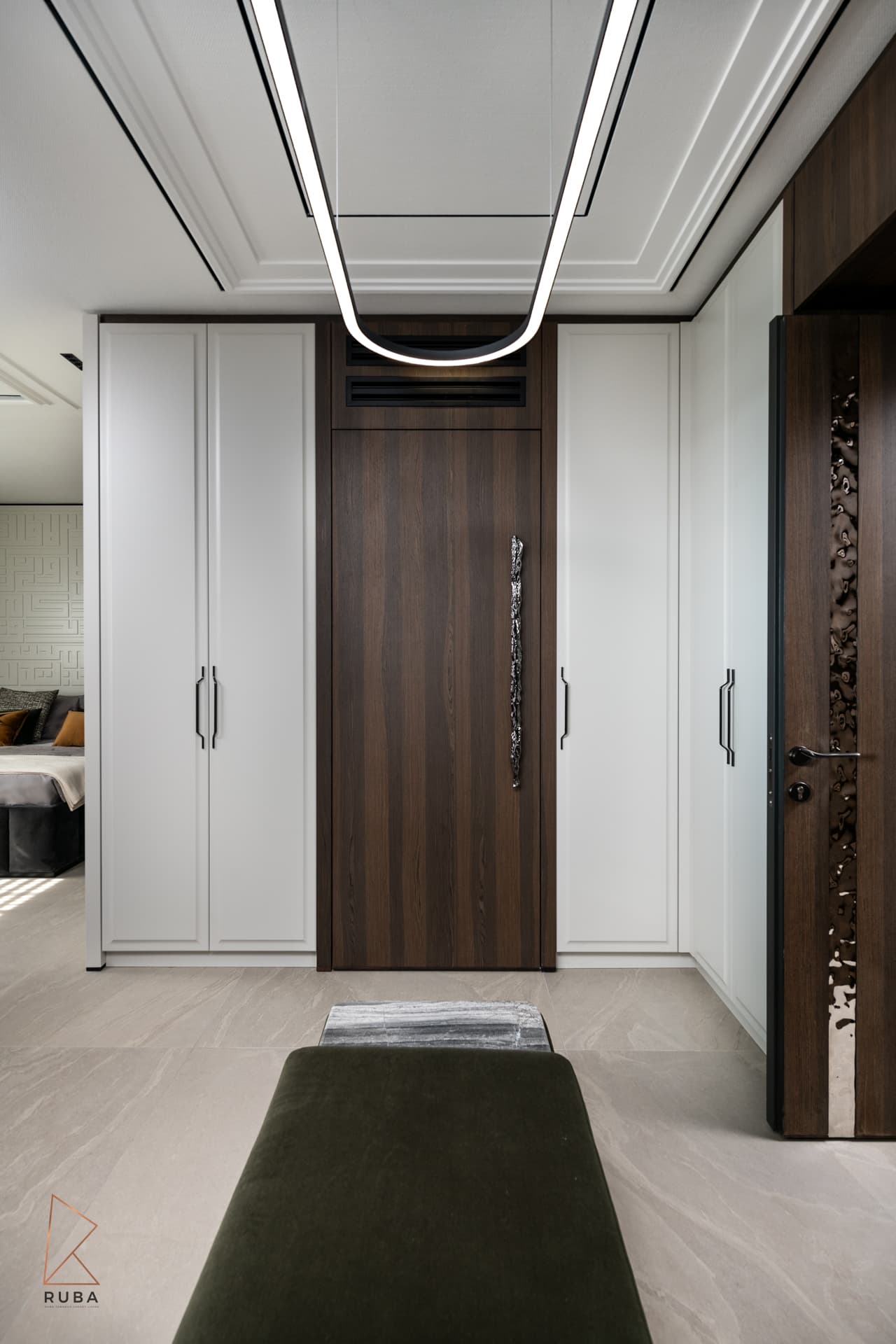 White wardrobes framing a walnut door with a sculpted pull under an oval ring light, seen past a velvet bench in Khayyal