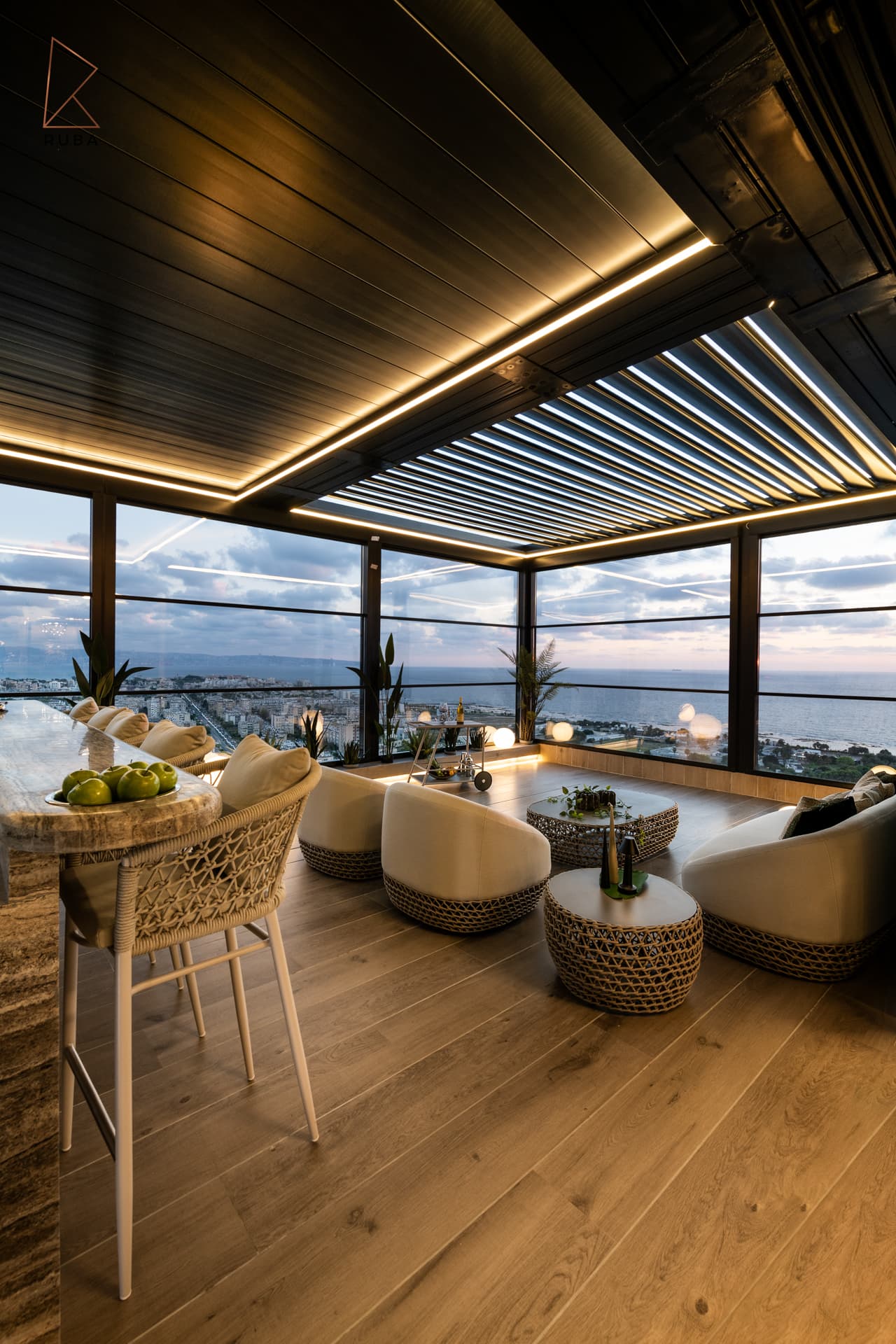 Glass-enclosed terrace lounge with woven armchairs, oak floor and a lit louvred pergola at sunset in Khayyal