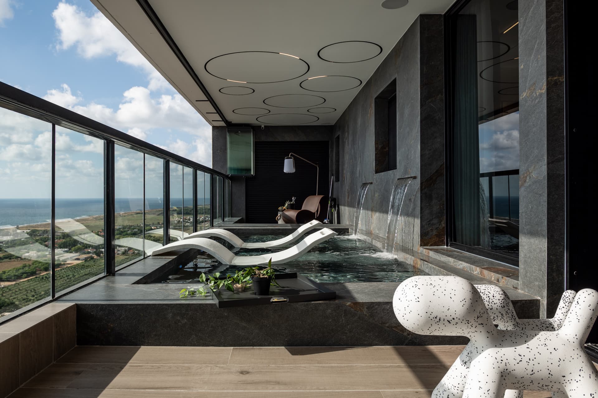 Long terrace pool with white in-water loungers, wall waterfalls and speckled dog-shaped seats beside a glass railing in Khayyal