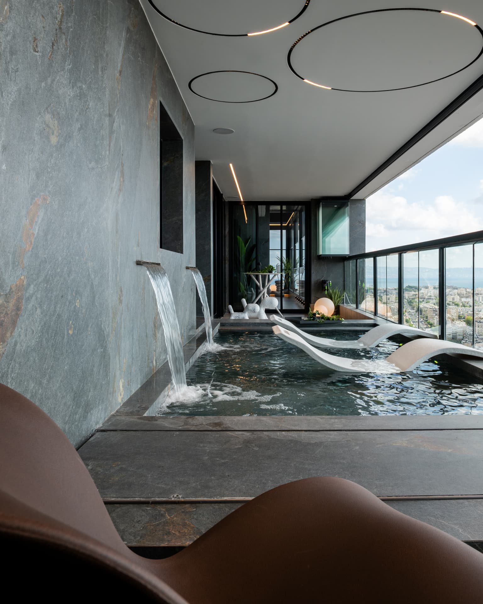 Twin waterfall spouts on a dark slate wall pouring into the terrace pool beside white loungers in Khayyal