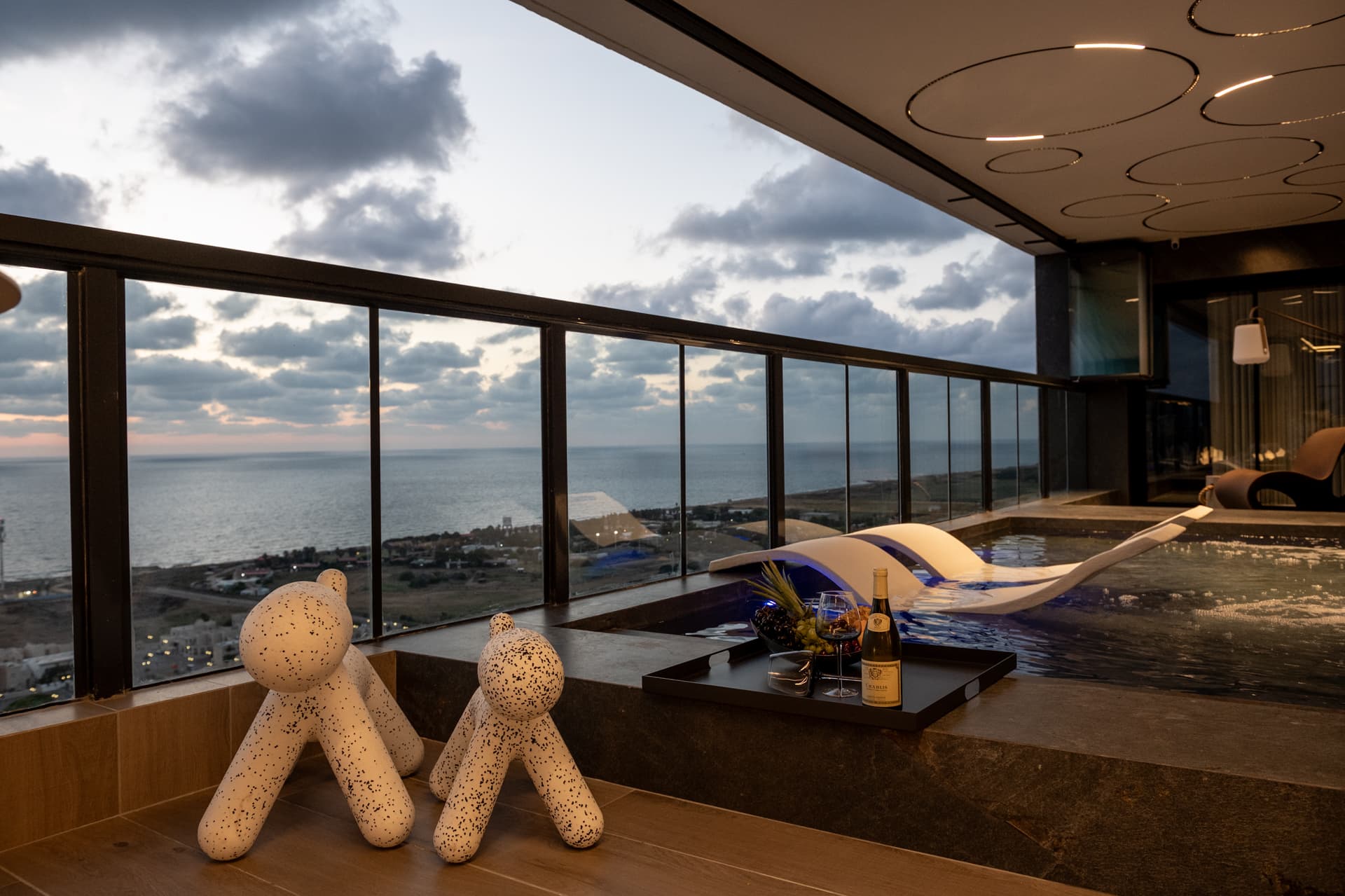Two speckled dog-shaped seats beside the pool edge with a wine tray and cloudy sea view at dusk in Khayyal