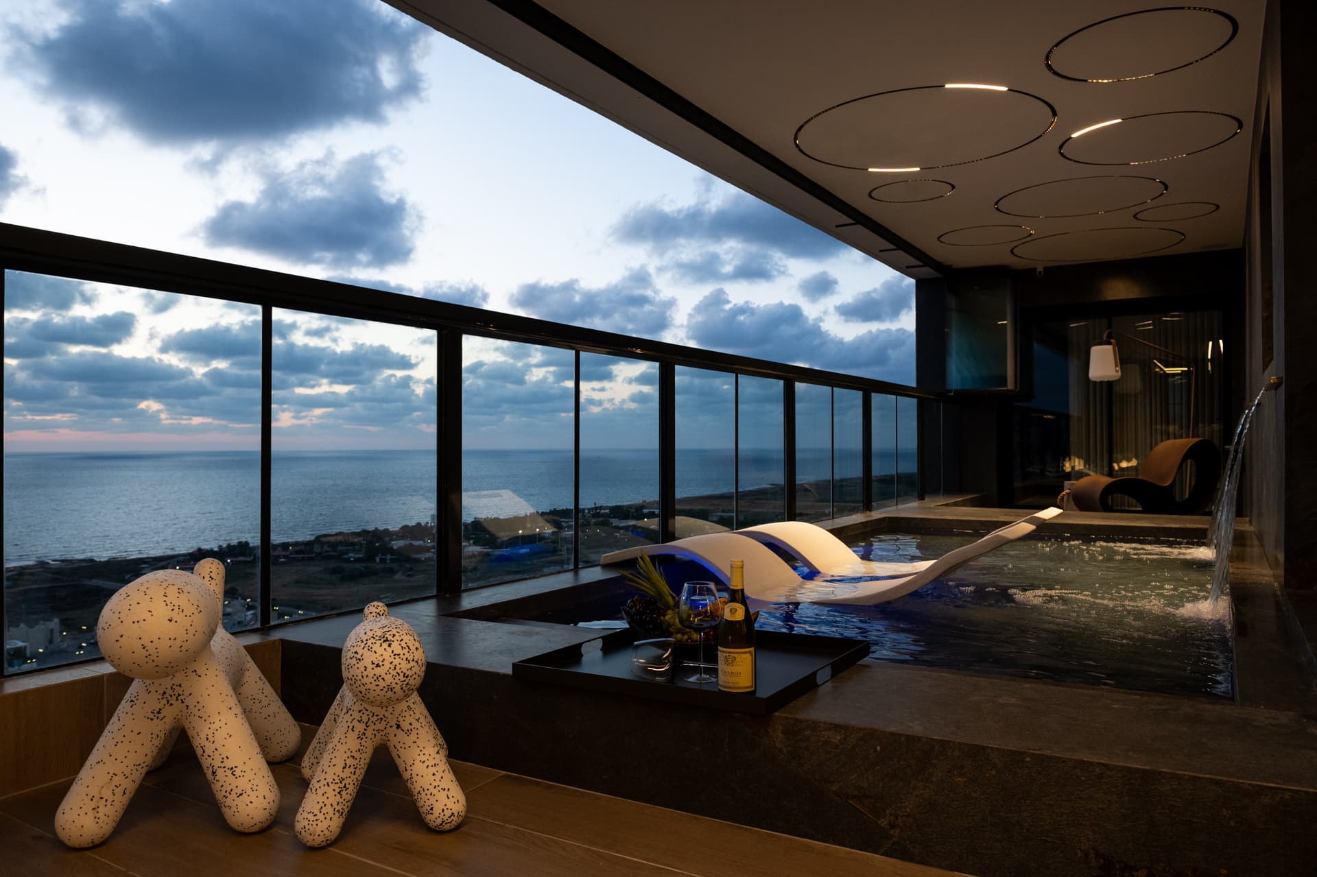 Dusk view of the pool with blue-lit loungers, a waterfall spout and dog-shaped seats under ring ceiling lights in Khayyal