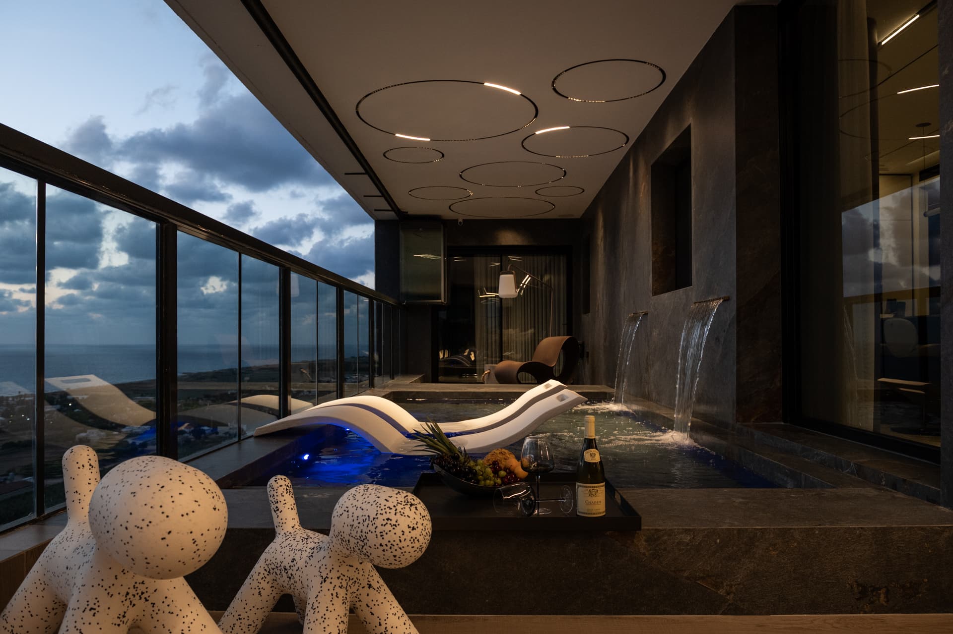 Evening pool terrace with blue underwater light, waterfall spouts and speckled dog-shaped seats in the foreground in Khayyal