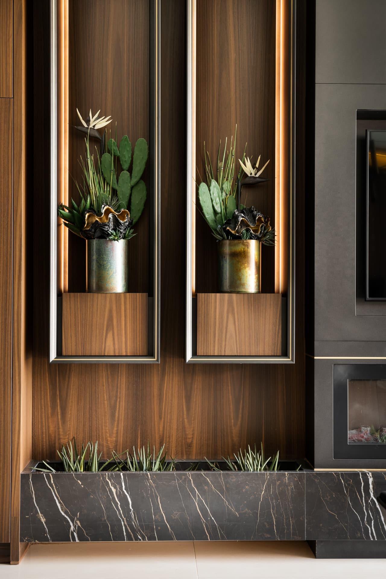 Two backlit walnut niches with patinated metal pots of cactus above a black veined stone planter, beside a fireplace, in King David