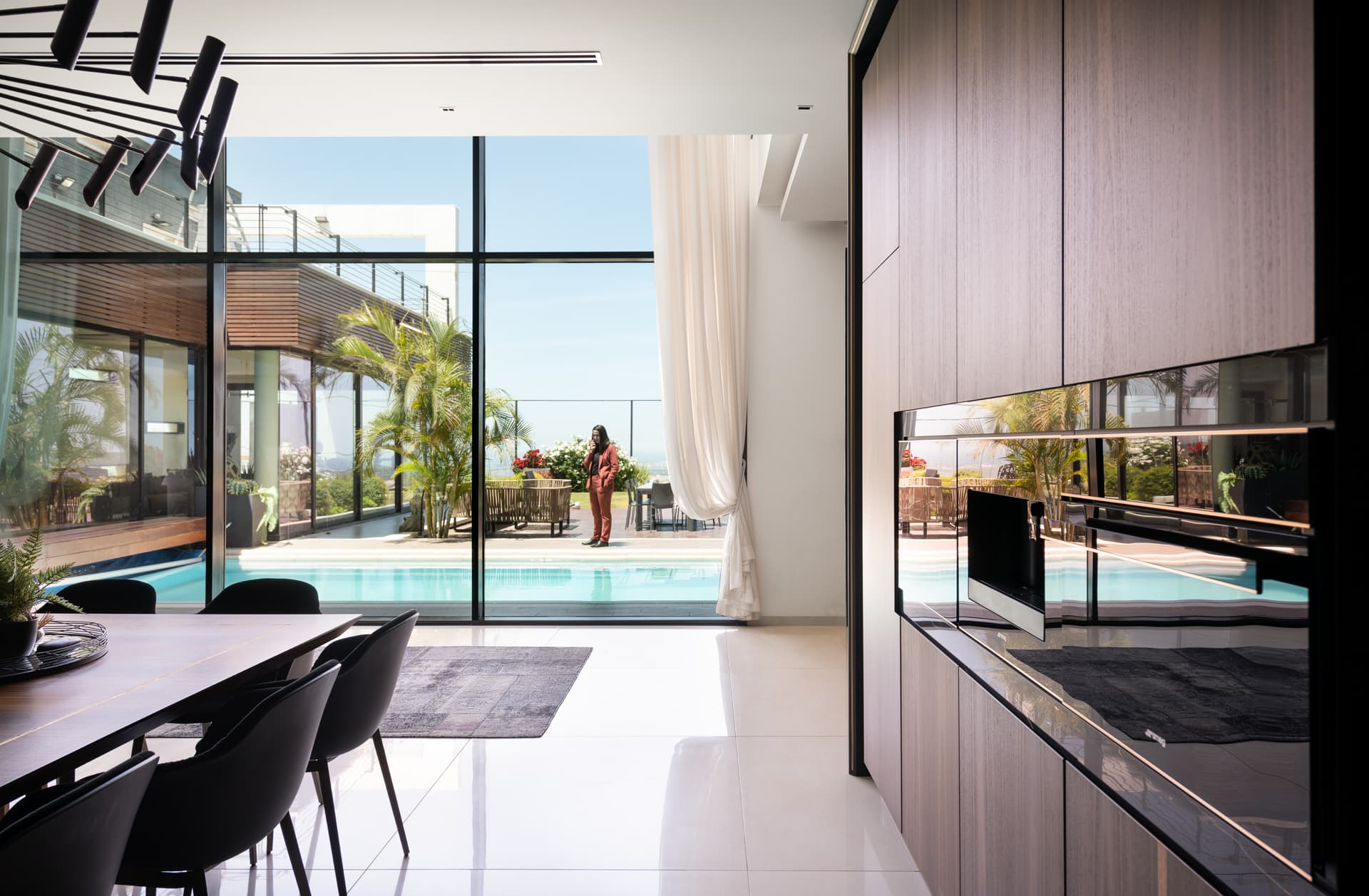 Dark wood cabinet wall with mirrored black appliances beside a dining table and full-height glazing onto the pool in the kitchen of King David