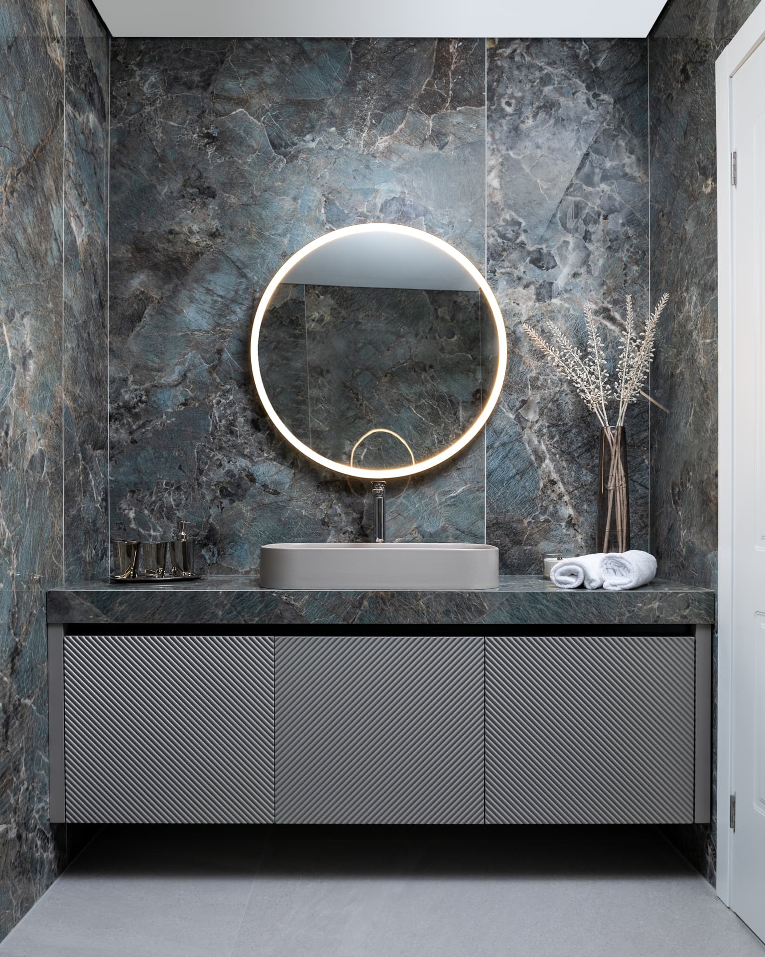 Backlit round mirror over a floating vanity with ribbed grey doors against blue-green veined stone in the bathroom of Marquina