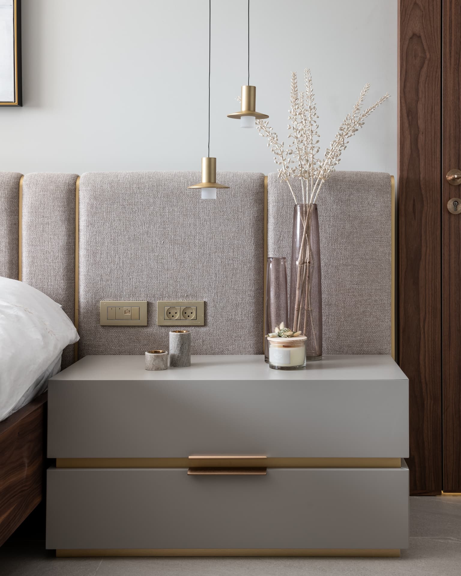 Grey bedside chest with a brass pull under twin brass pendants against the upholstered headboard in the bedroom of Marquina