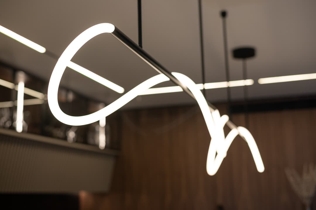Glowing looped LED tube pendant on black rods with linear ceiling lights and glass cabinets blurred behind in the kitchen of Marquina