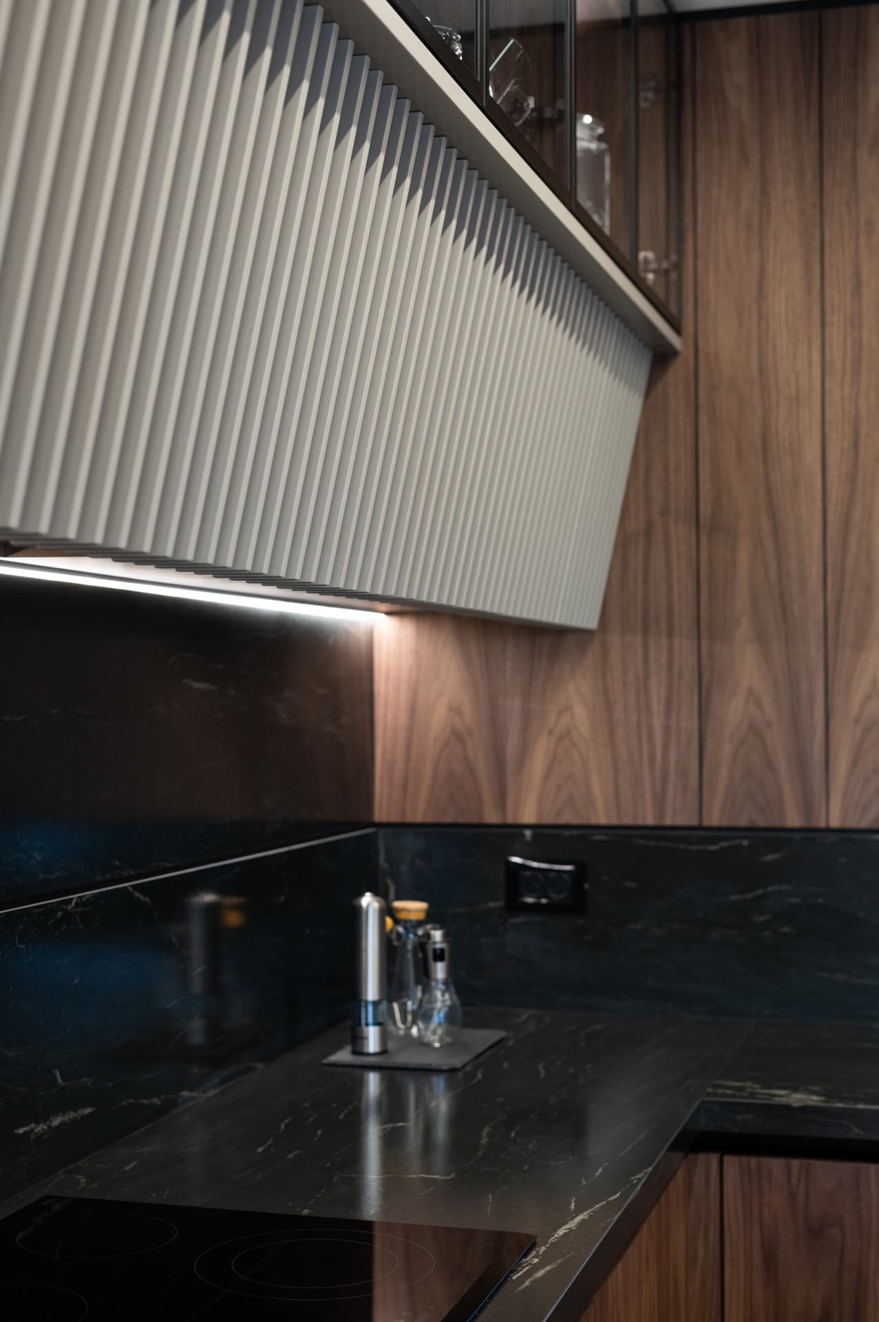 Angled fluted pale hood panel lit from beneath, above a black veined stone backsplash and cooktop in the kitchen of Marquina