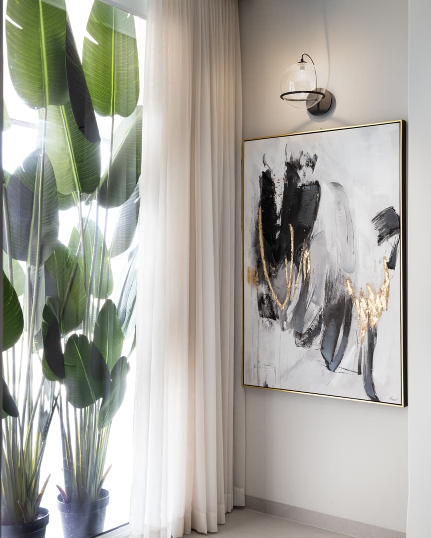 Abstract black and gold painting under a glass globe wall light beside sheer curtains and tall leafy plants in Marquina