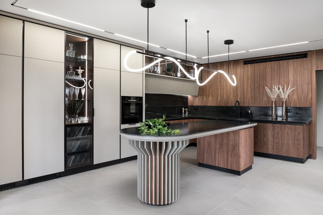 Rounded island on a fluted pedestal under sculptural LED pendants, with a lit glass display cabinet in the kitchen of Marquina